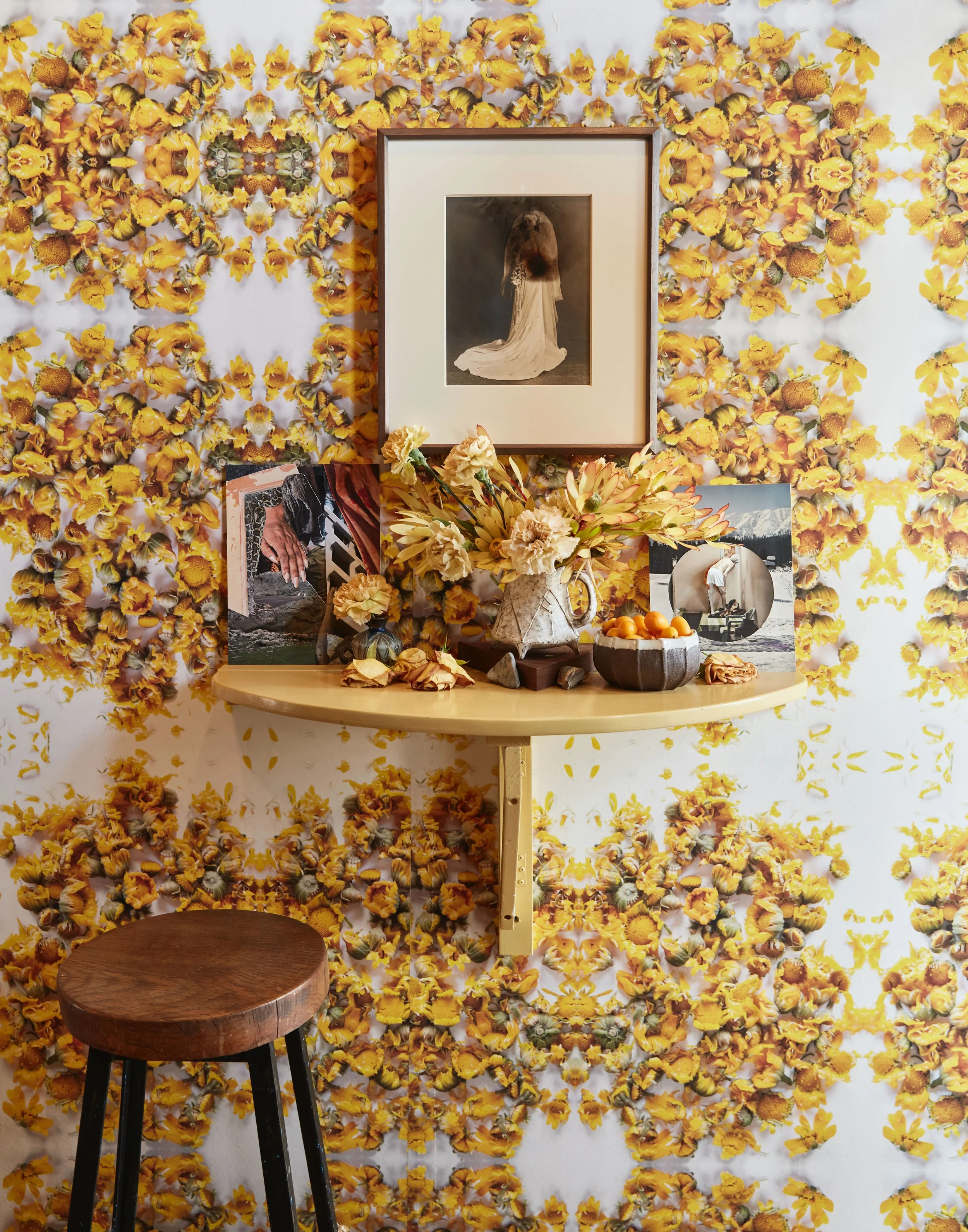 Decorative space featuring a wall with yellow floral wallpaper, a mounted framed photograph of a woman in a white dress, a small yellow shelf with dried flowers in a vase, and photos of a person by a river and mountains, with a wooden stool in the fo