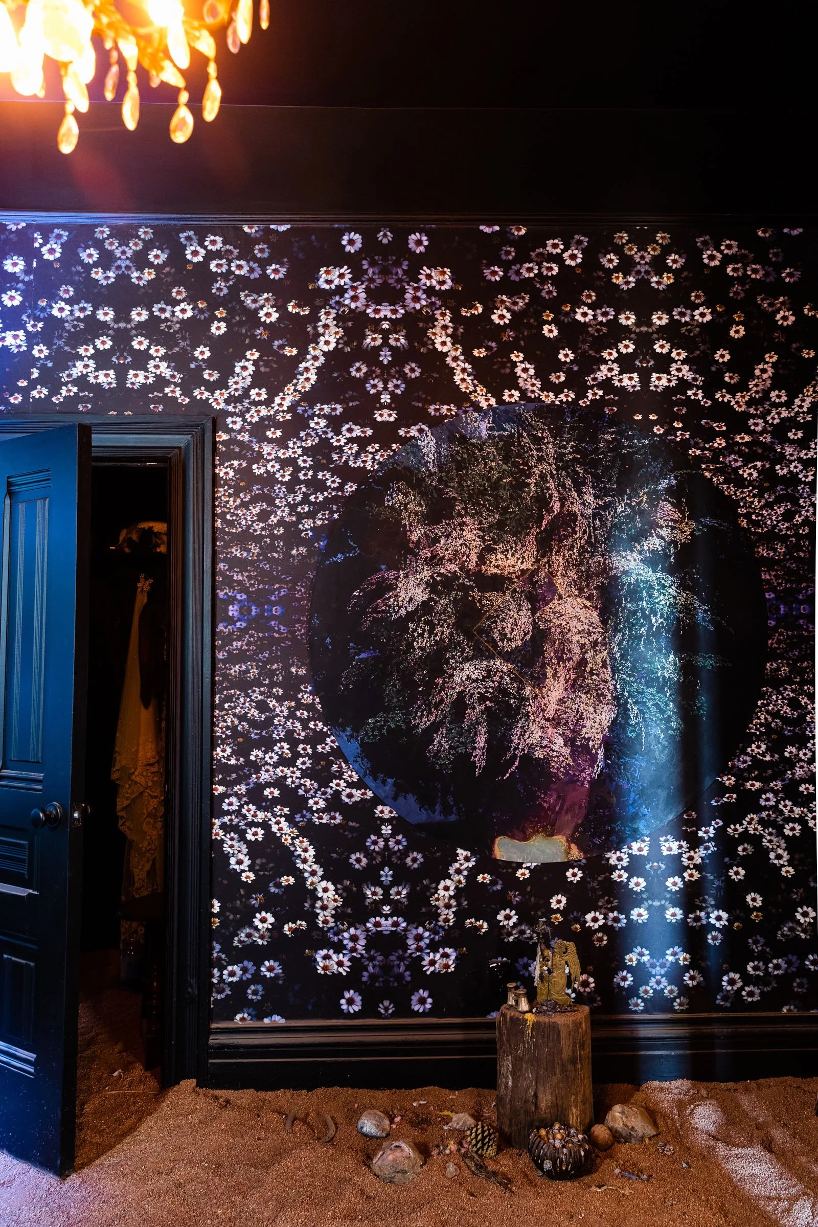 Interior wall with floral theme, large round art piece of trees, star-shaped text reading 'NATURAL', a door partially open revealing clothing, wooden log with small statue, and rocks on sandy floor.