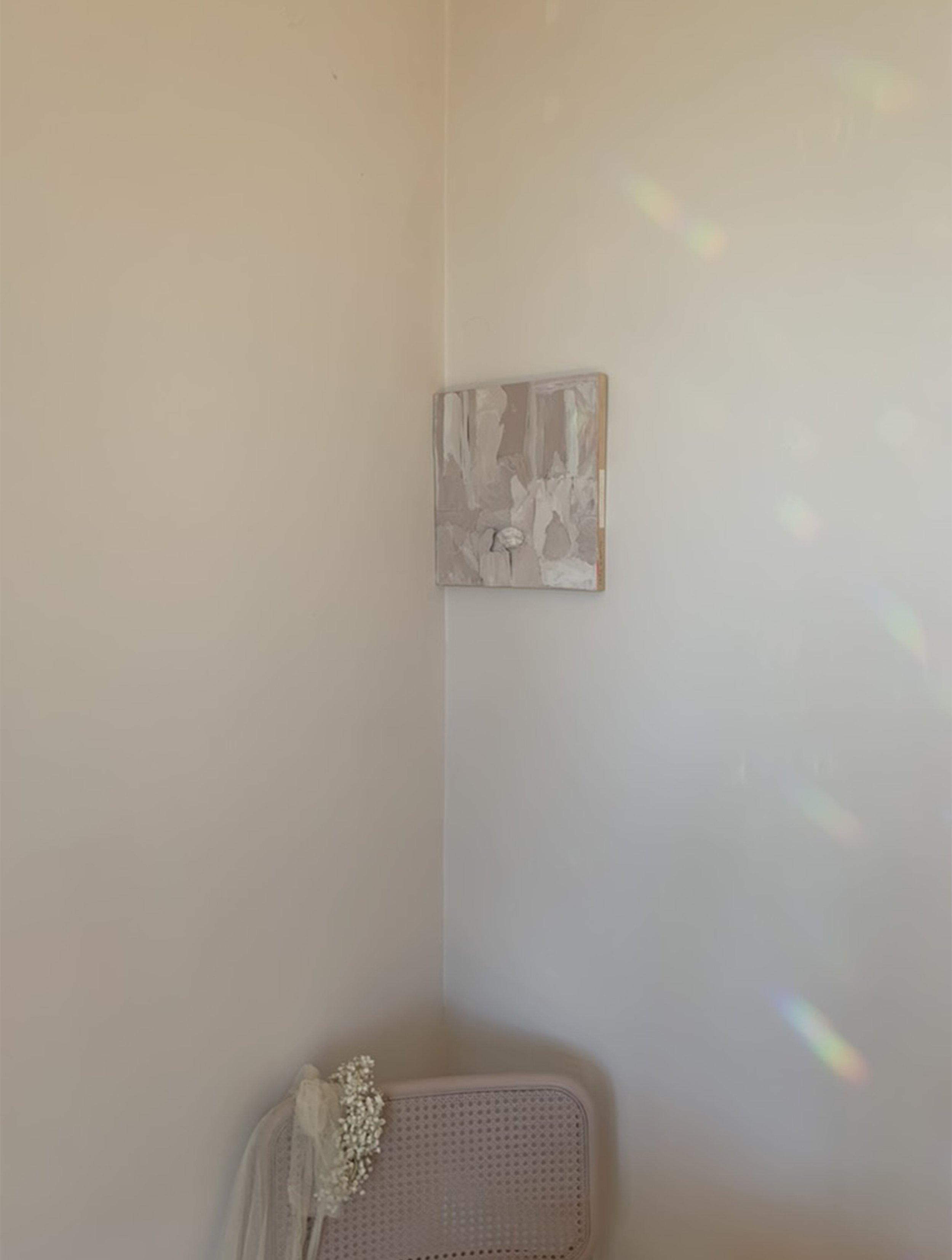 A corner of a room with cream-colored walls, a small abstract painting on the wall, and a pink headboard with a bouquet of white flowers resting on it.