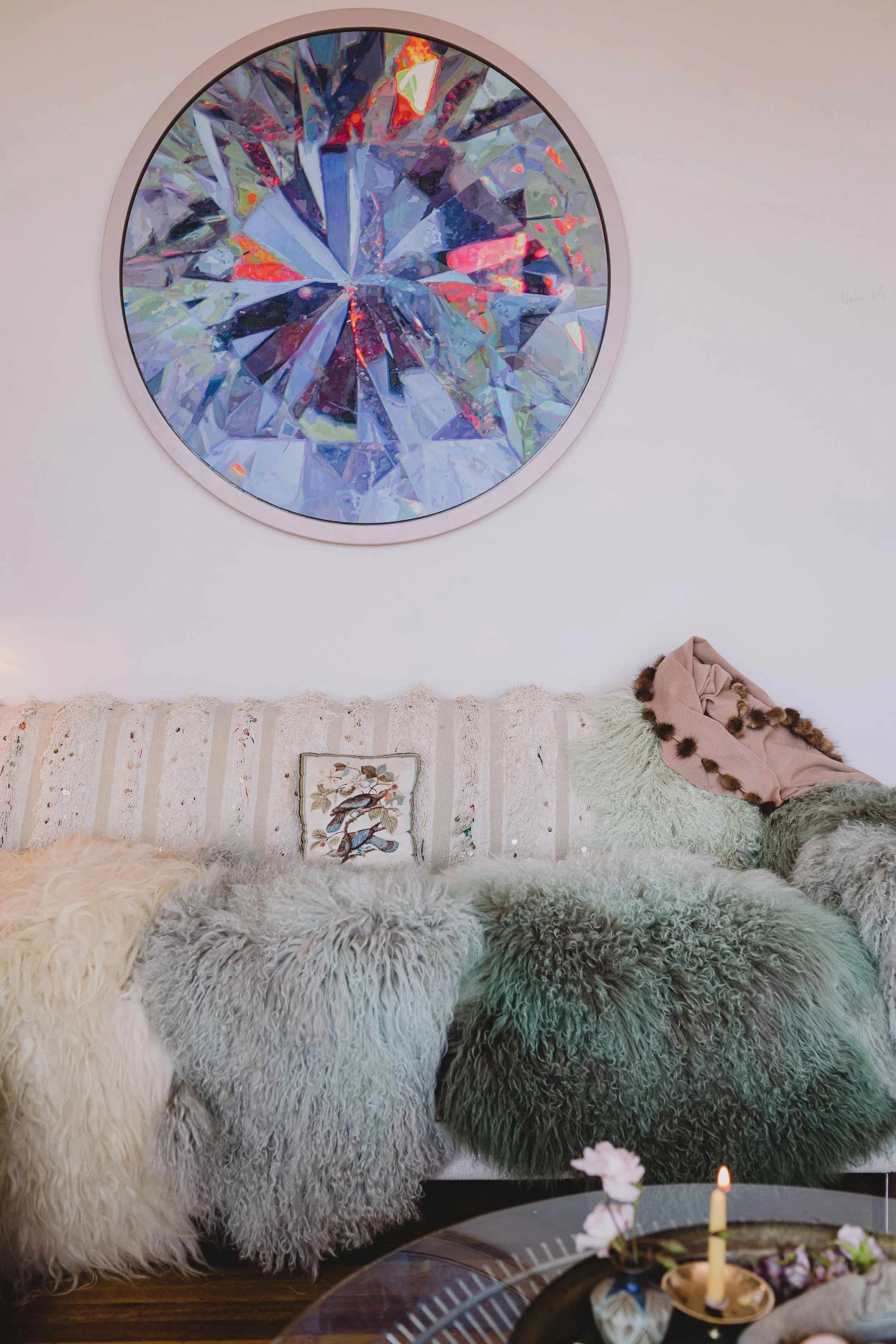 Living room with a large round mirror resembling a diamond on the wall, cream-colored striped sofa with various fuzzy pillows and throws, a small embroidered pillow with bird design, and a coffee table with a candle and flower arrangement.