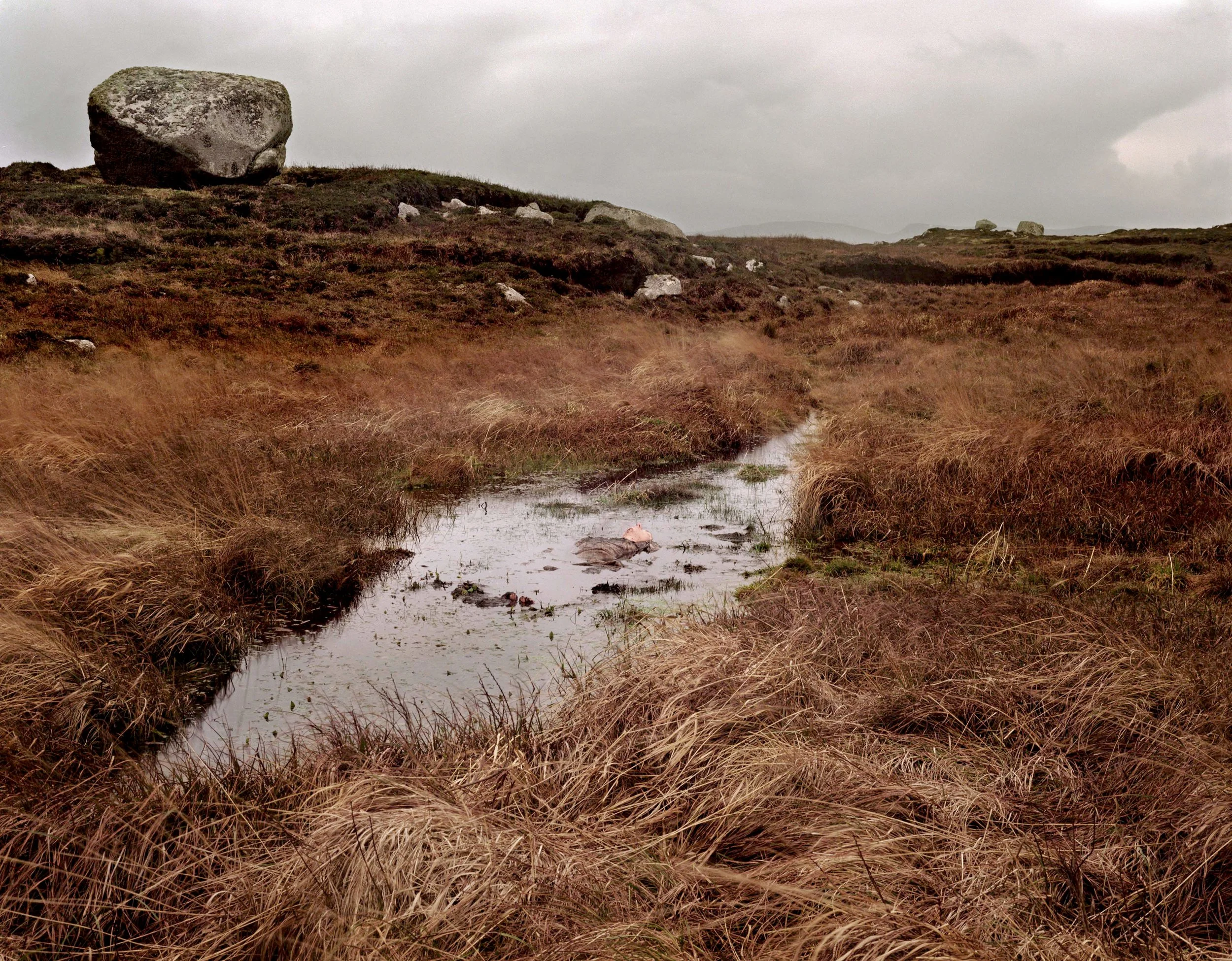 A small stream running through brown grassy moorland with large rocks, overcast sky, and cloudy weather.