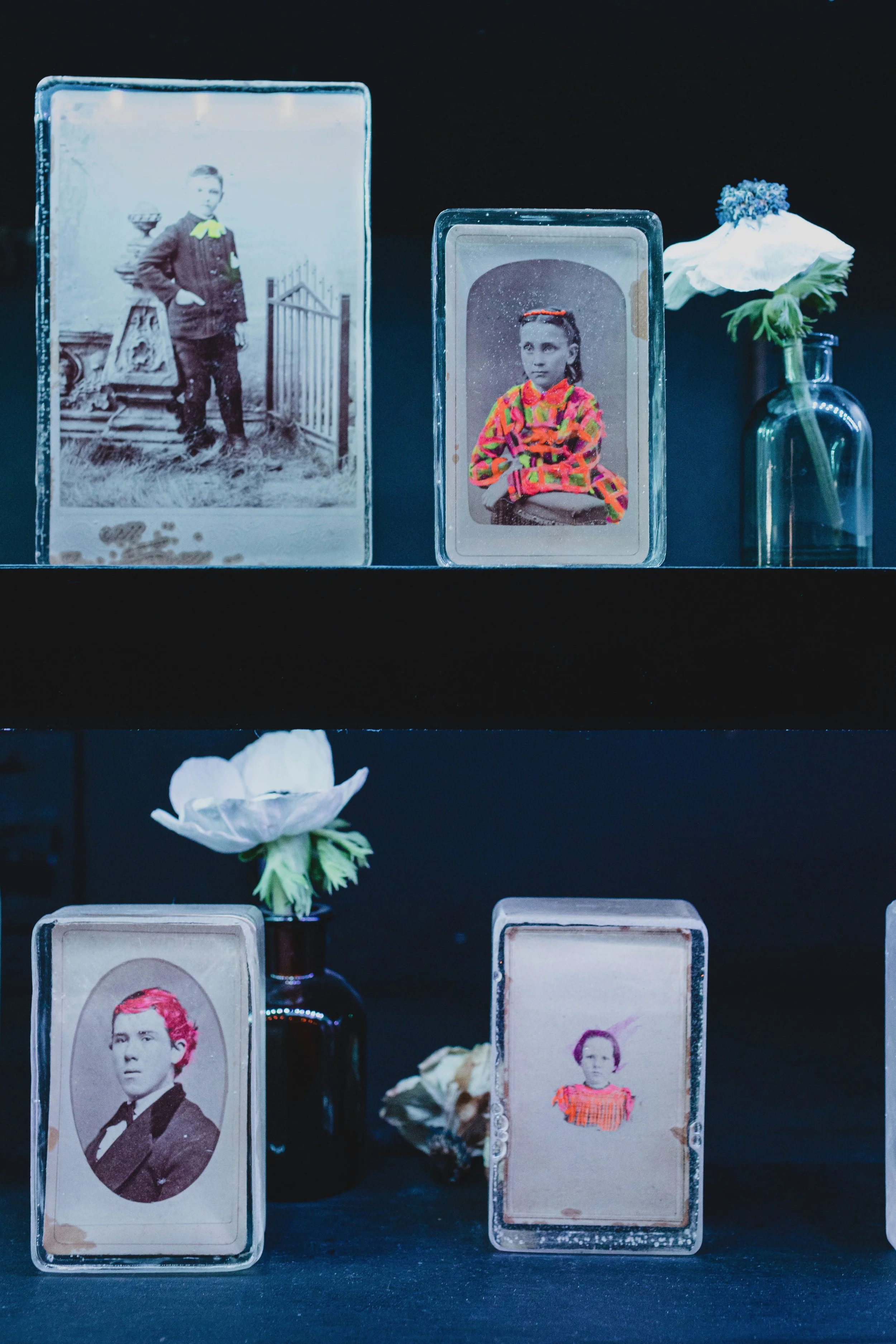Display of five vintage photographs of children, three on top shelf and two on bottom, with white and purple flowers in glass vases beside them, all set against a dark background.