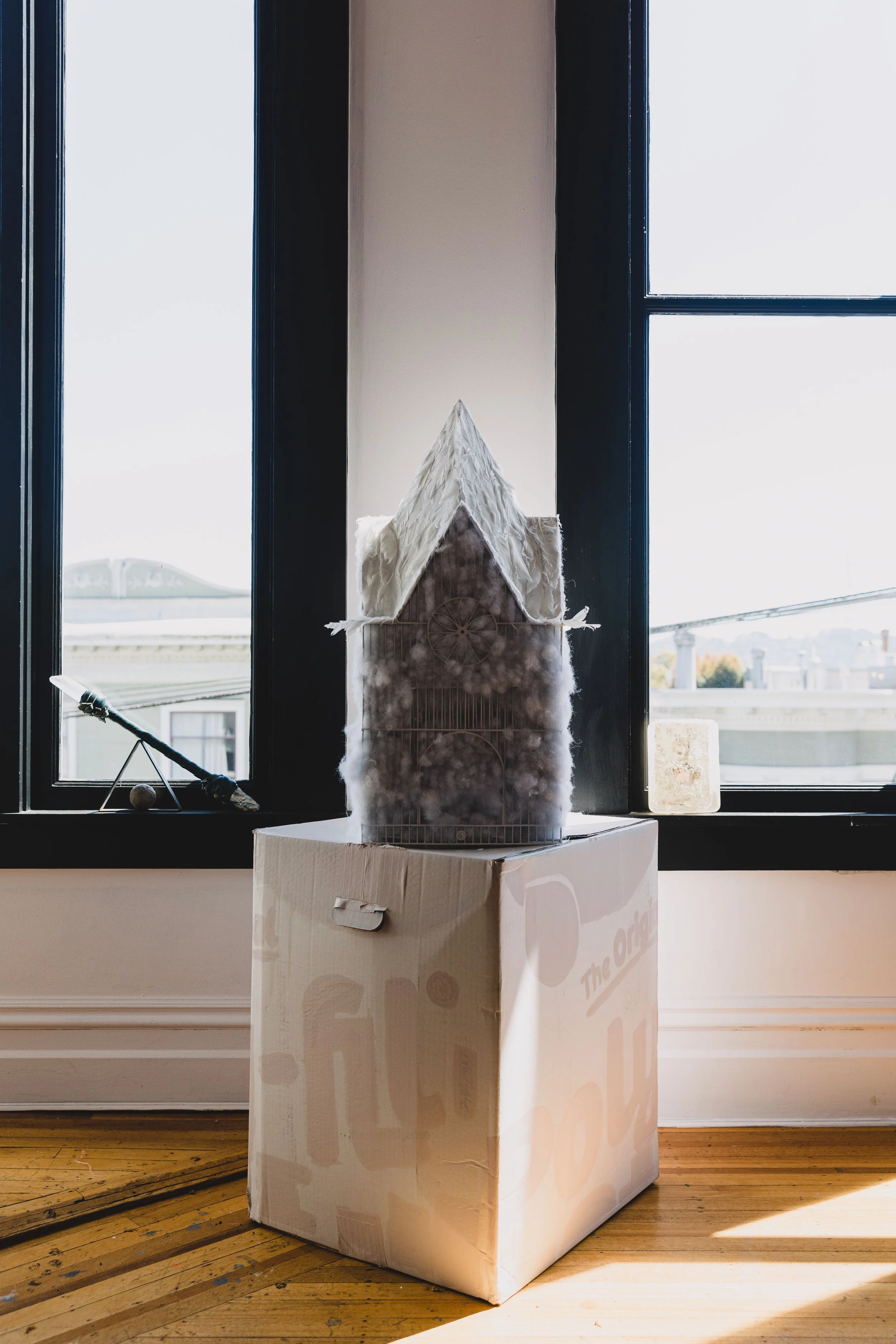 A model of a house with a steep roof, placed on a cardboard box, positioned in front of two large windows in a room with wooden flooring.