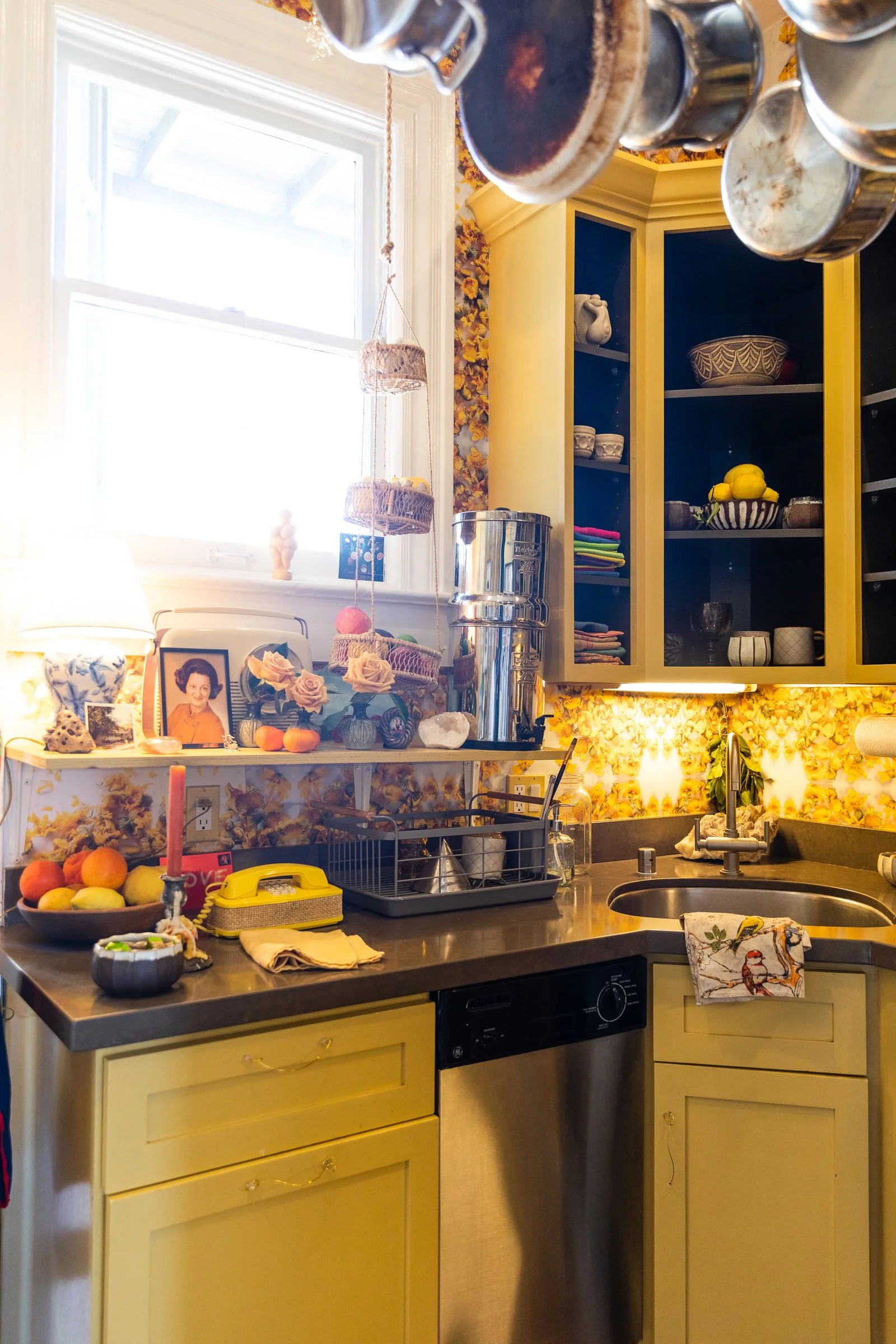 A colorful kitchen with yellow cabinets, a stainless steel dishwasher, and a window with sunlight. The countertop has various items including a bowl of fruit, a framed photo, flowers, a dish rack, and a candle. Open shelves and a cabinet display dish