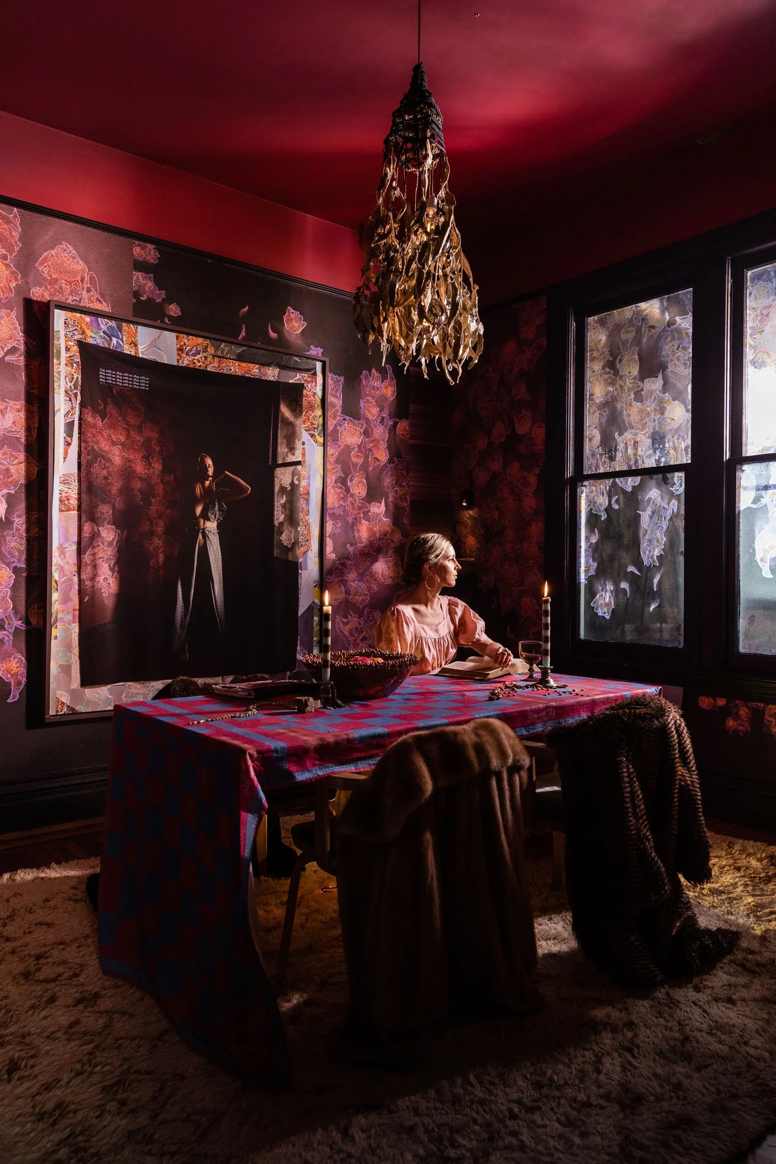 A woman sitting at a dining table in a dimly lit room with pink and purple floral wallpaper, a chandelier hanging from the ceiling, and large windows with abstract designs on the glass.