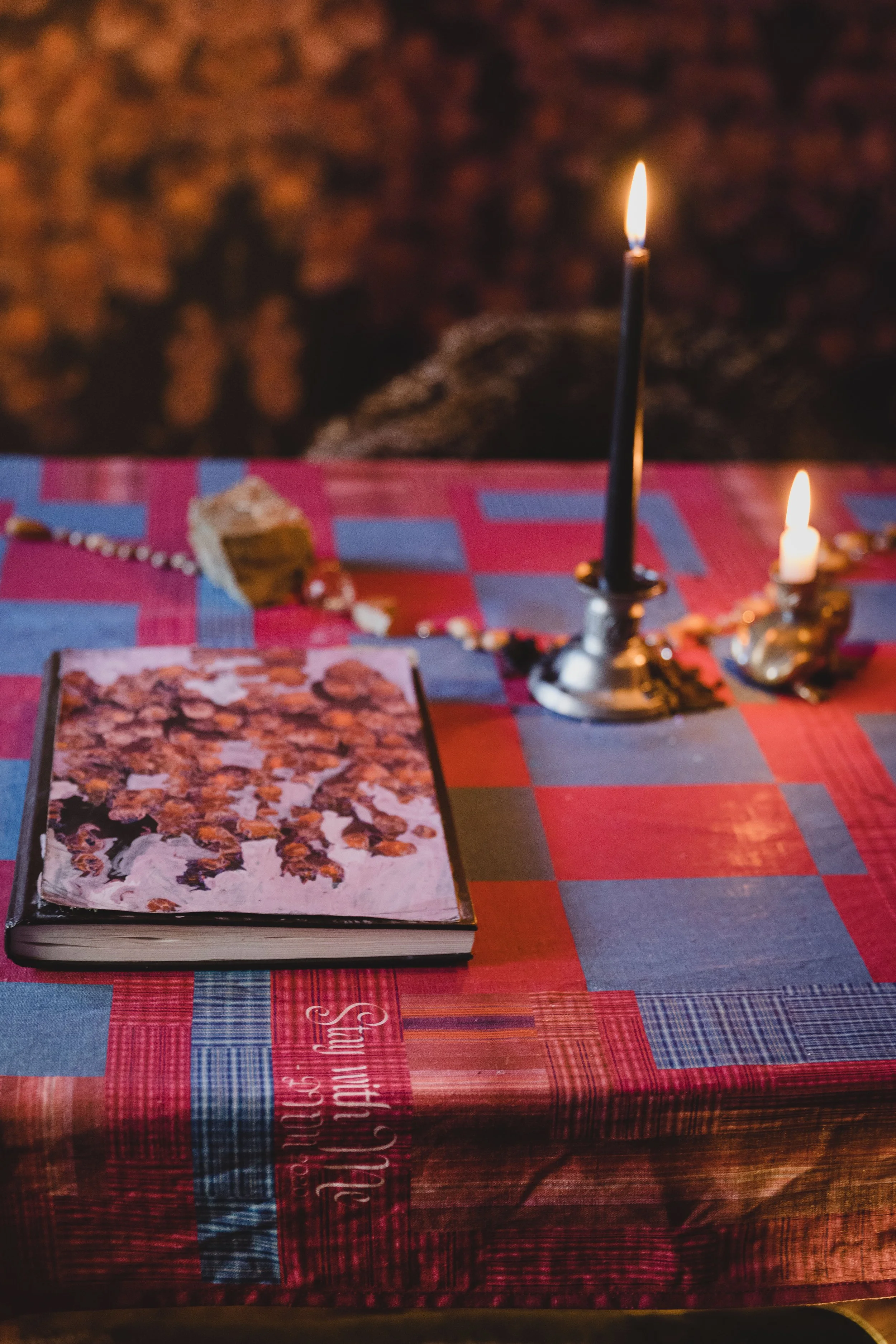 A table covered with a red and blue plaid tablecloth, holding an open book with a painting of sunflowers, a lit black candle in a silver holder, and a beaded necklace or rosary, with blurred warm background.