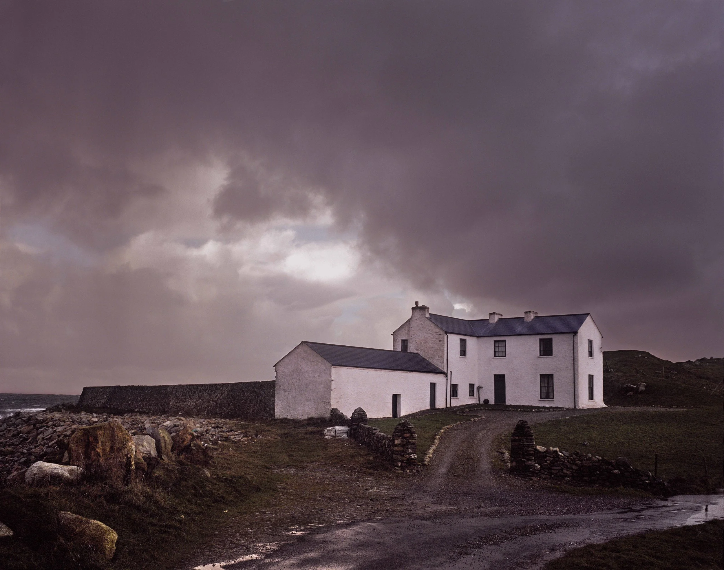 A white house with dark roof, surrounded by rocky yard and a stone wall, under a dark, cloudy sky.