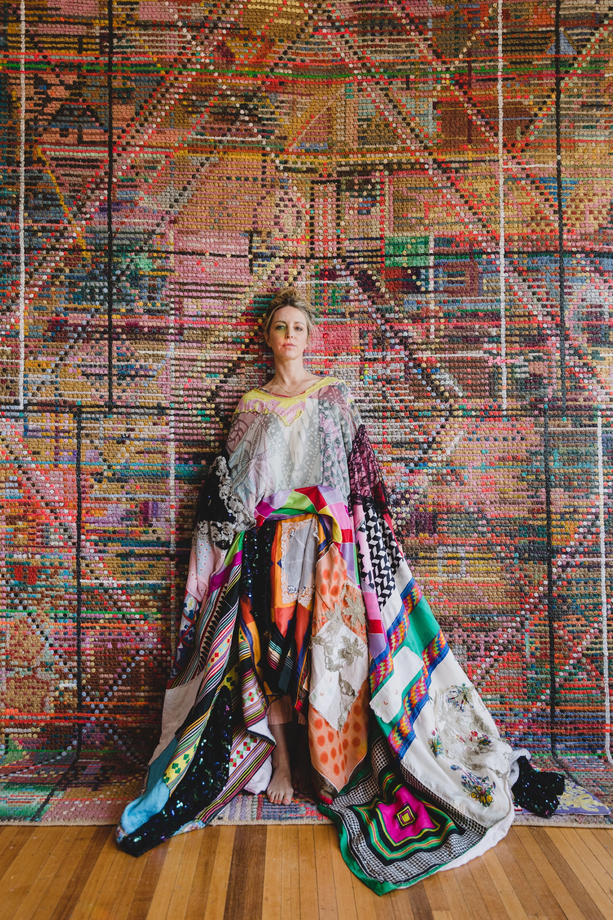 Woman standing barefoot in front of a colorful, intricate mosaic wall, wearing a patchwork dress made of various fabrics and patterns.