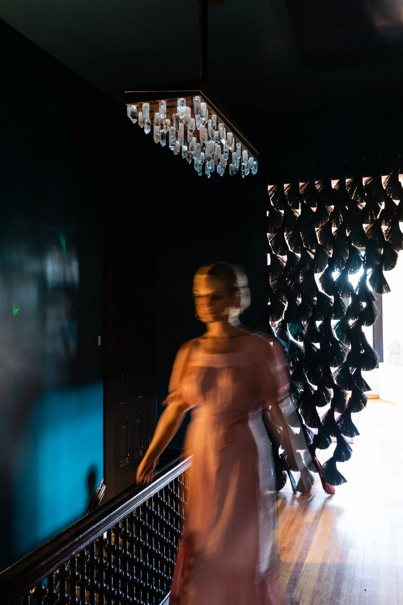 A woman in a light pink dress walking indoors near a black wall with a decorative curtain and a chandelier hanging from the ceiling.