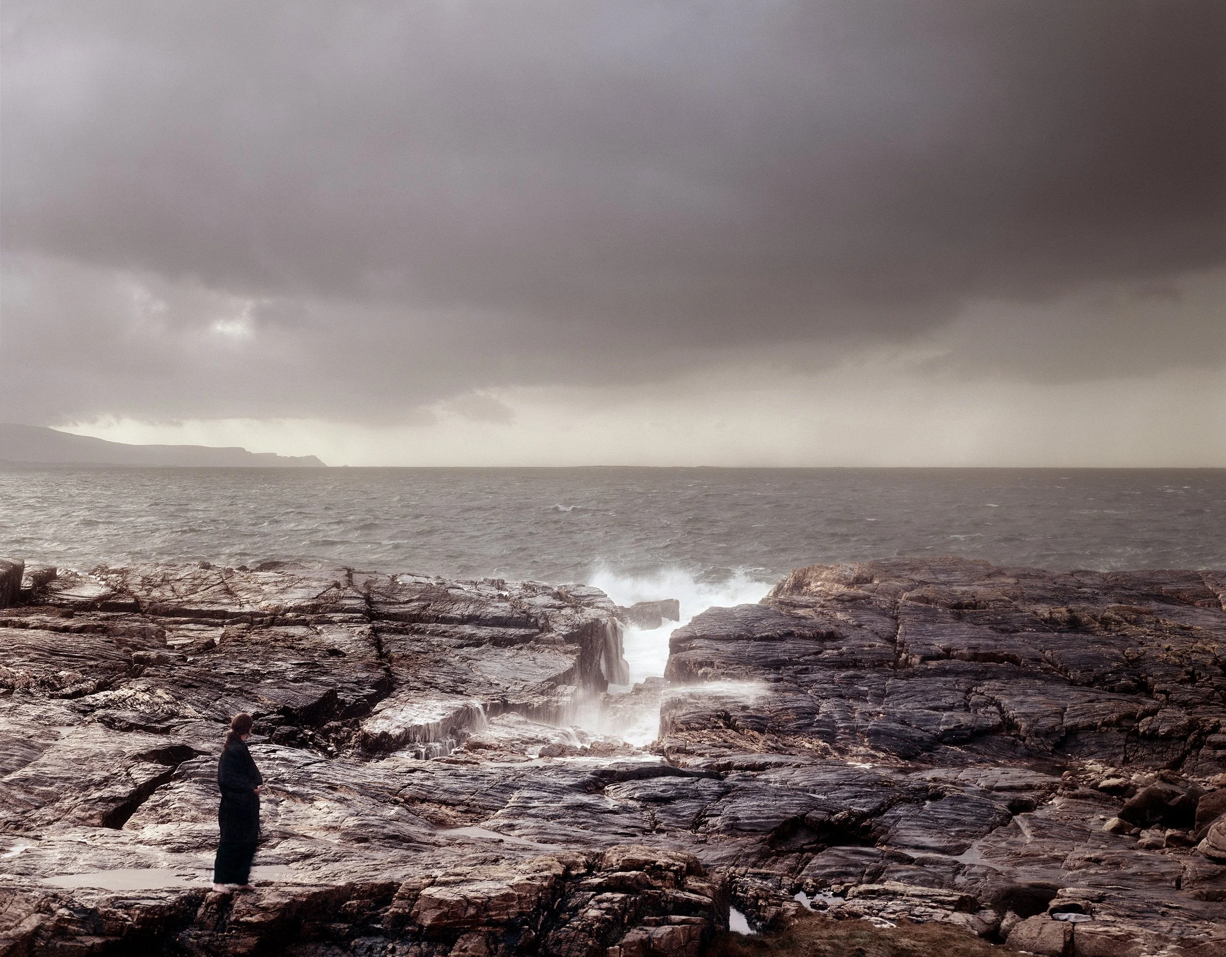 Person standing on rocky coastline under dark, stormy clouds with waves crashing against rocks.