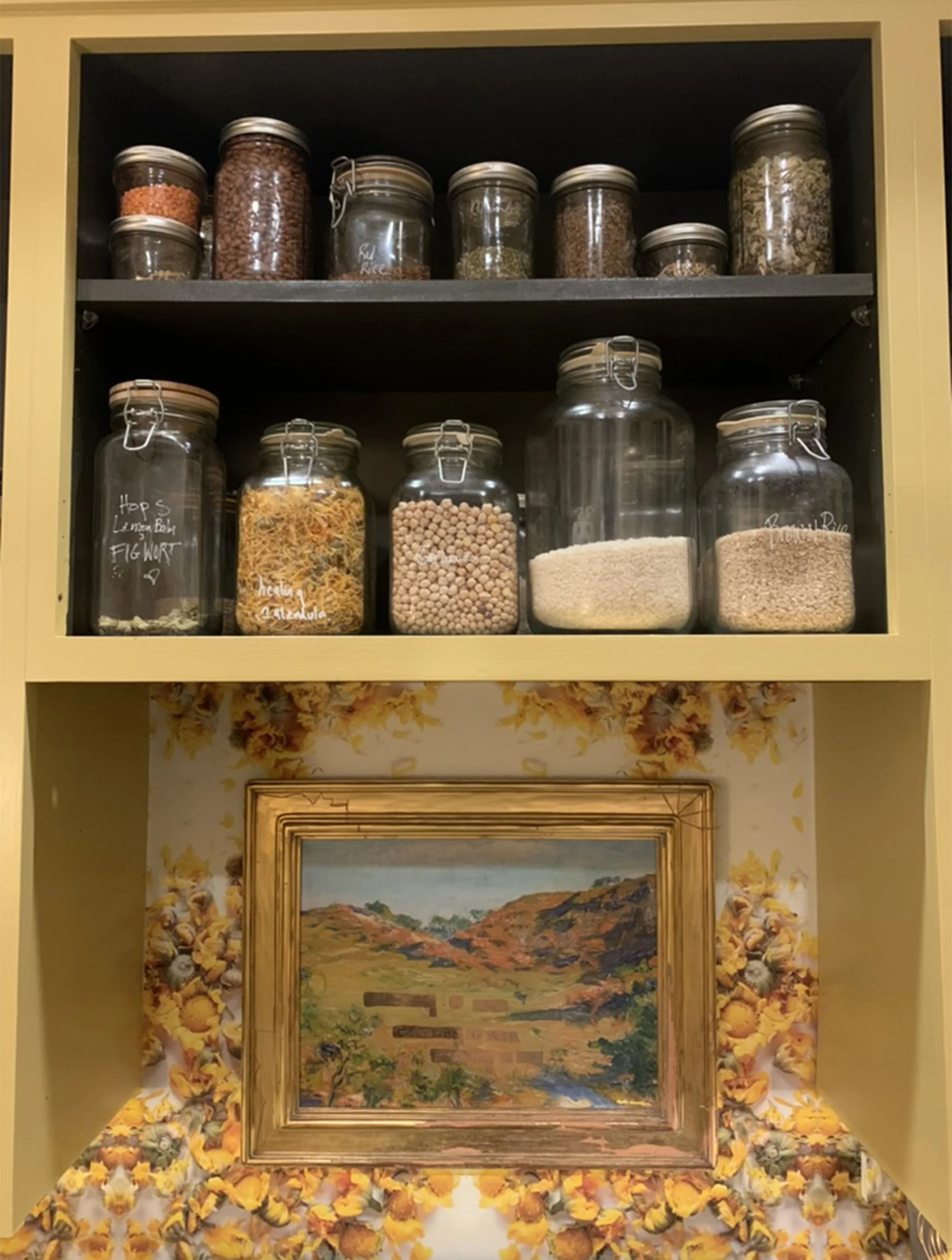 Kitchen cabinet with glass jars containing spices and grains, above a framed landscape painting surrounded by sunflower and fall leaves decor.