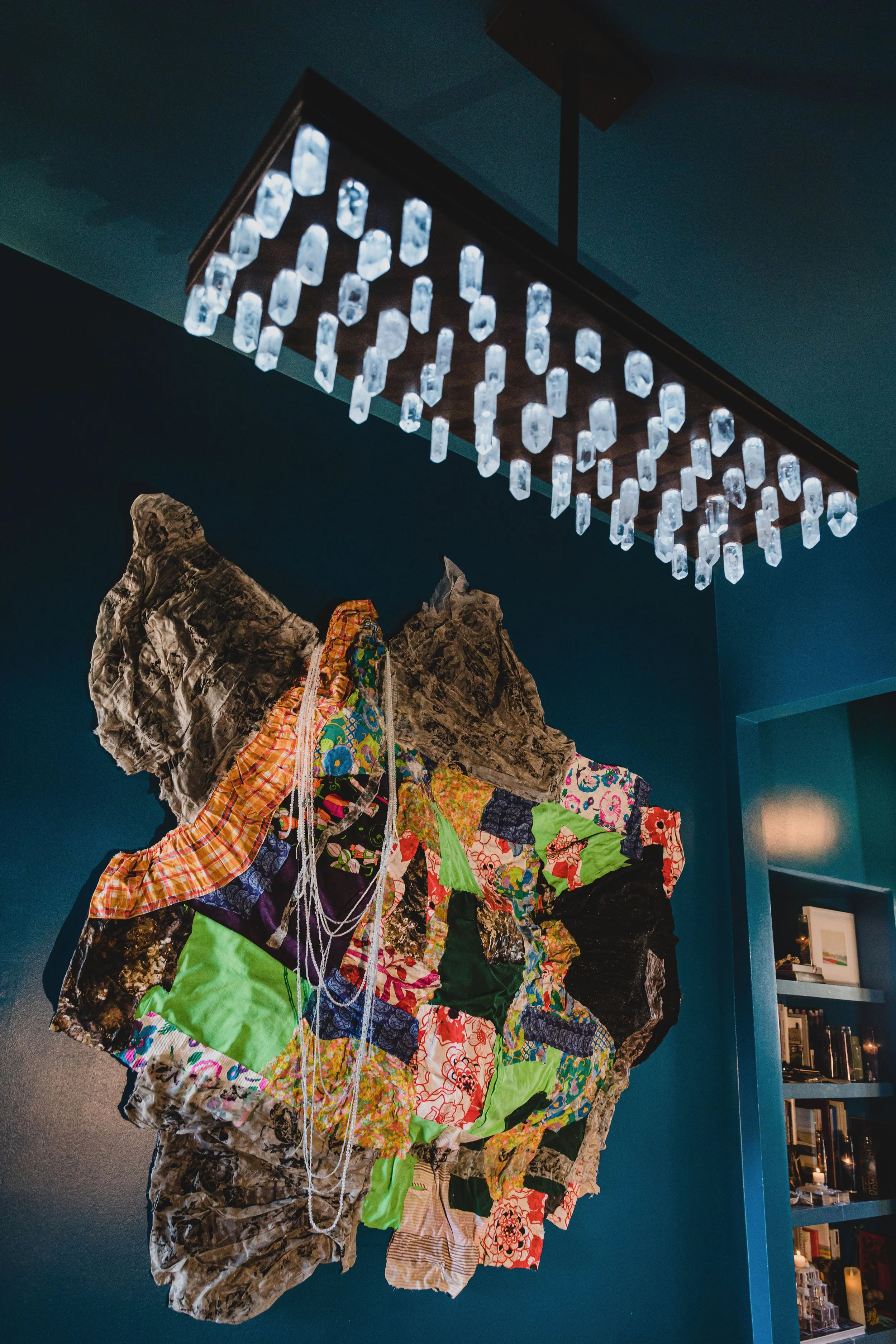 Art installation featuring a large, irregularly shaped collage made of various colorful fabric pieces and textured materials, mounted on a dark blue wall, illuminated by a modern ceiling light fixture with multiple clear, rectangular lights.