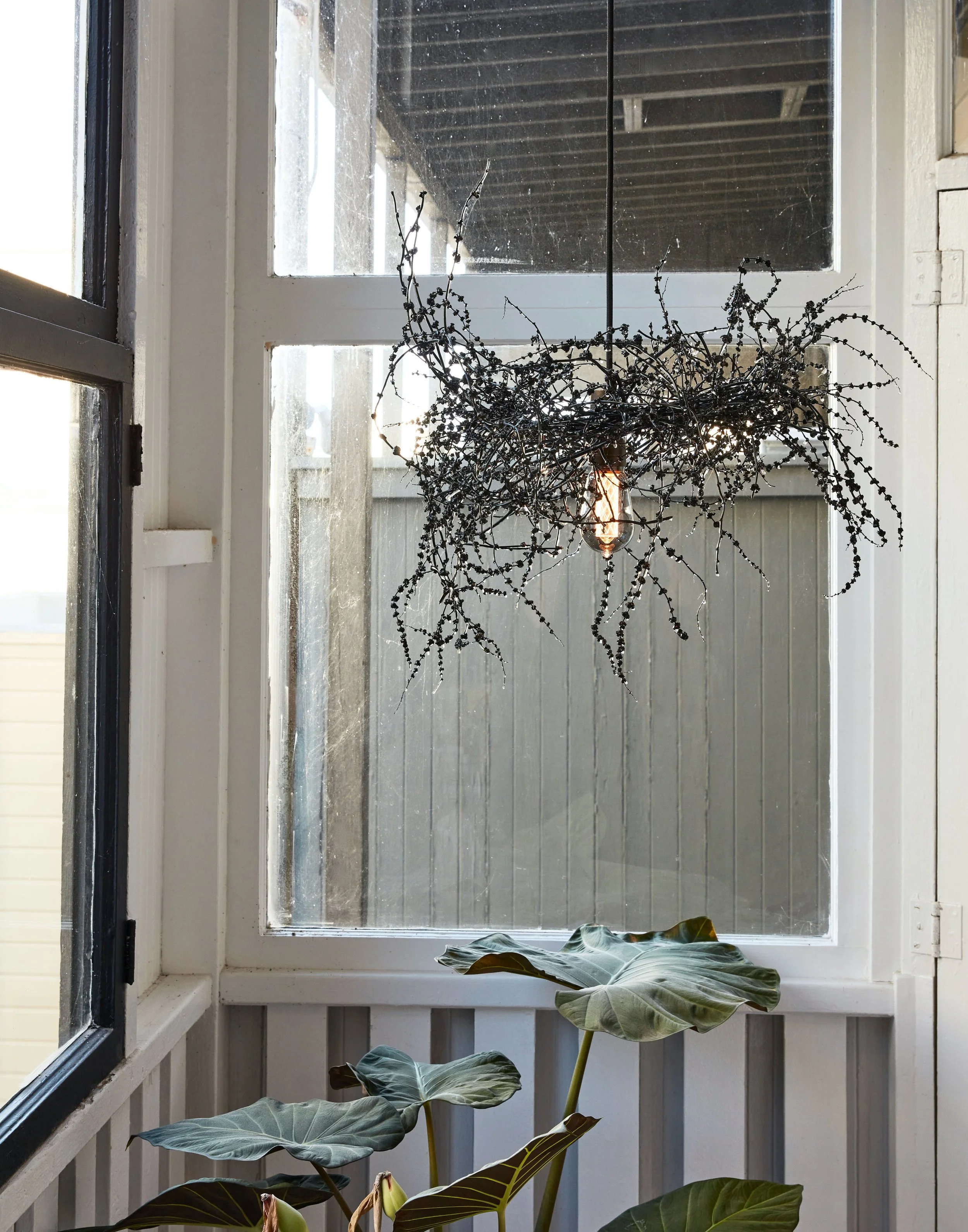 Decorative hanging light fixture made of black beads and branches, hanging in front of a window with small green plants below.