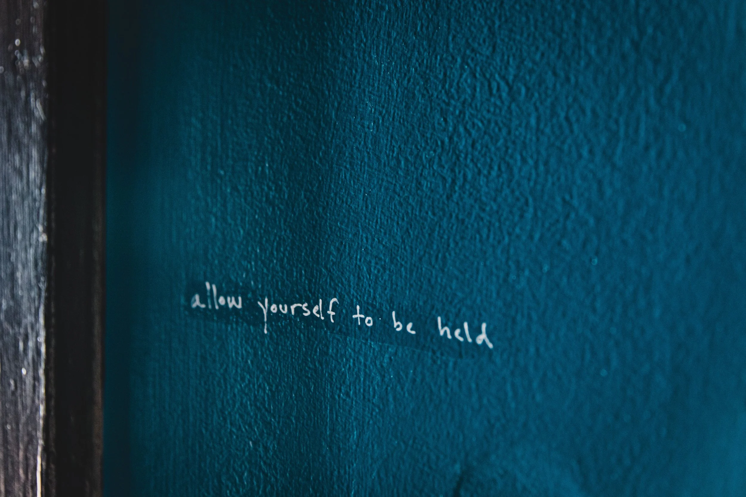 Close-up of a textured blue wall with white handwritten text that says, 'allow yourself to be held'.