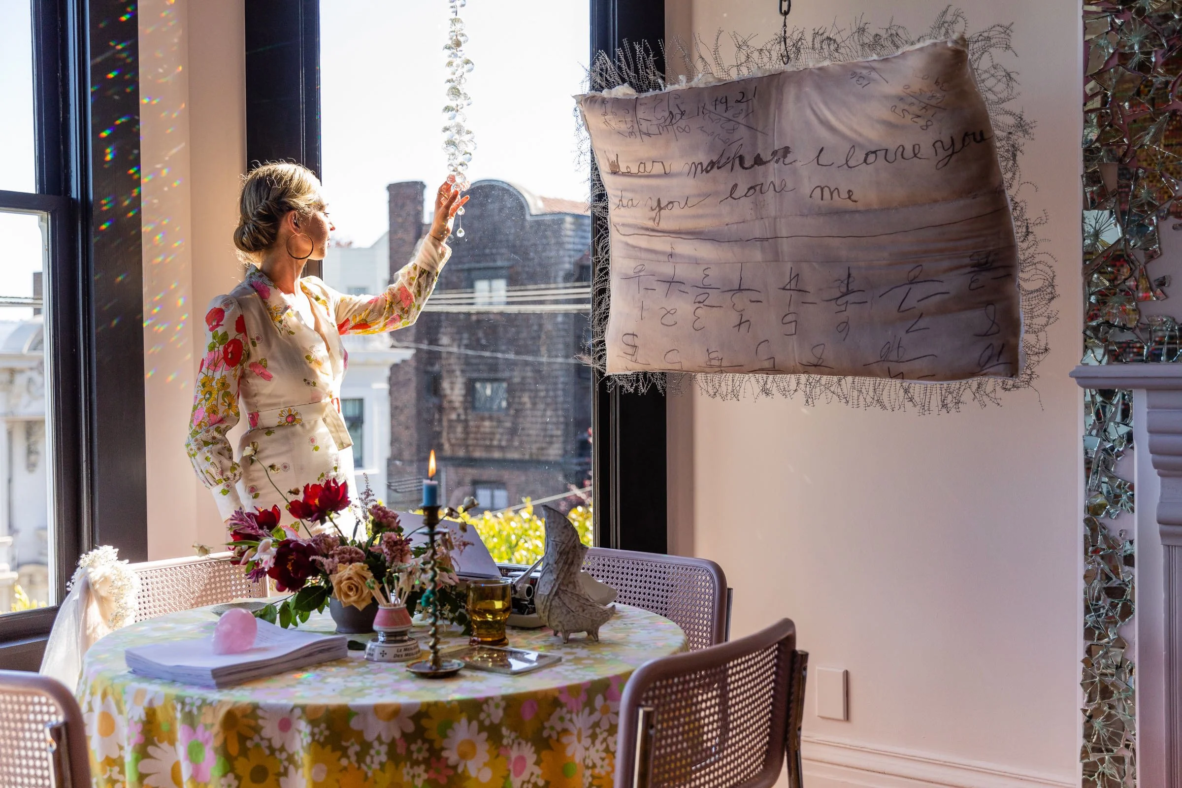 A woman in a floral dress standing at a window, holding up a string of crystals, with sunlight streaming in. A table with a floral cloth, flowers, a candelabra, a candle, and books is in the foreground. A large, fabric sheet with handwritten text and