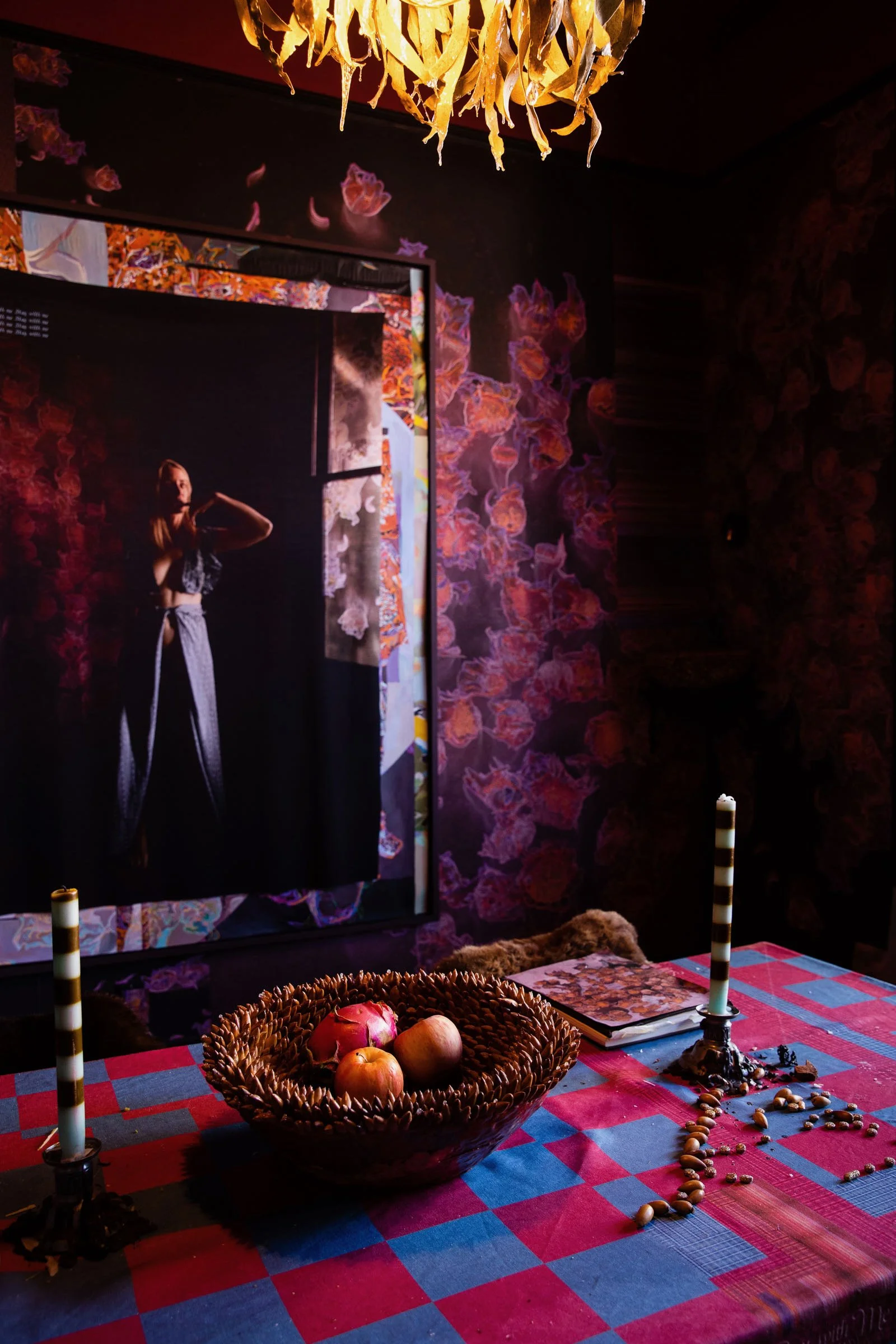 A decorated table with a woven basket of apples and pomegranate, and two candlesticks, in front of a dark wall covered with floral wallpaper and a mirror reflecting a woman in a black and white outfit.