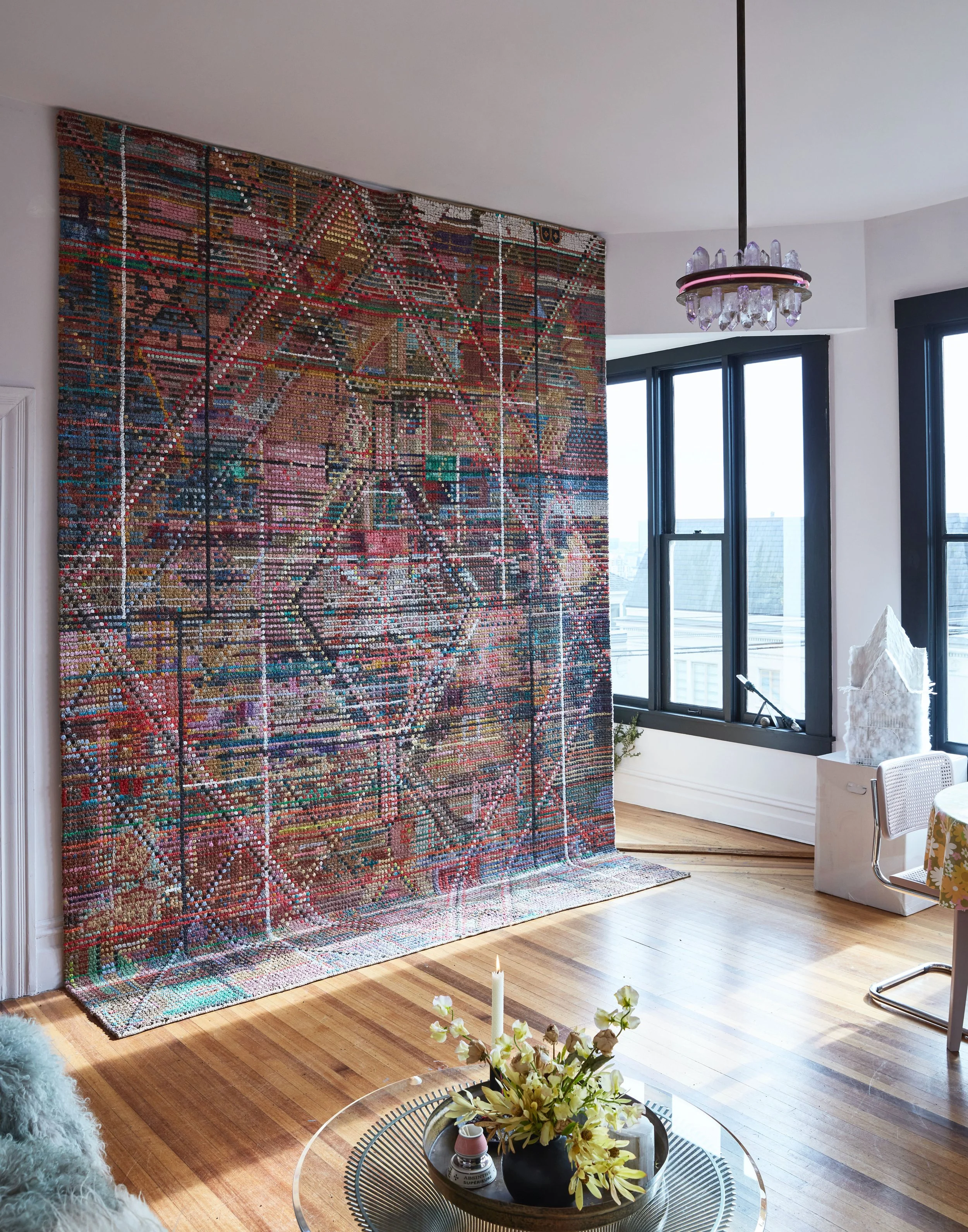 Colorful, abstract art installation hanging on a white wall in a modern apartment, with large windows, wooden floor, and a coffee table with flowers.