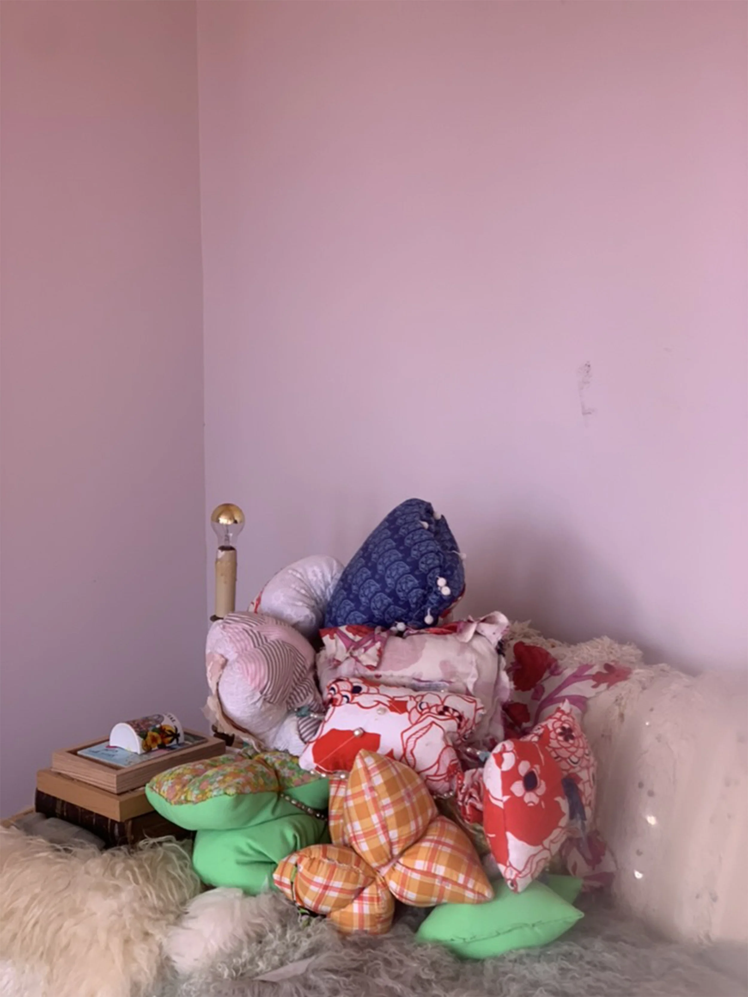 A pile of assorted decorative pillows in various patterns and colors, placed on a fluffy white rug, with a small wooden side table holding books and a sunflowers arrangement, against a plain light pink wall.