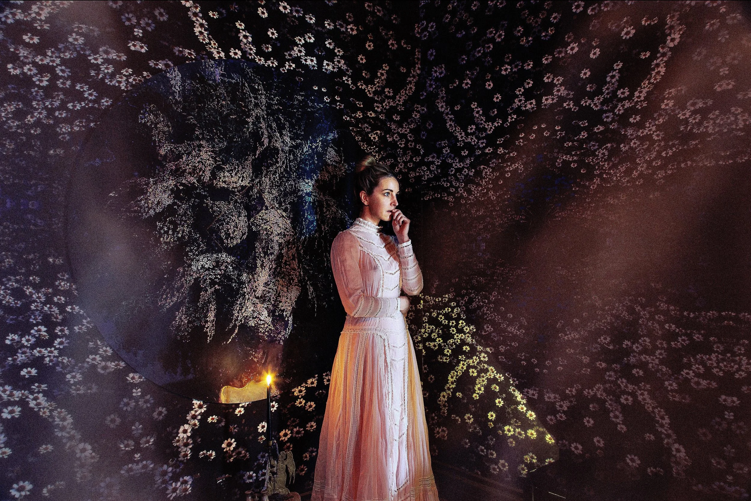 A woman in a pink, vintage-style dress stands in a dark room with intricate floral patterns on the walls and ceiling, illuminated by a small lit candle.