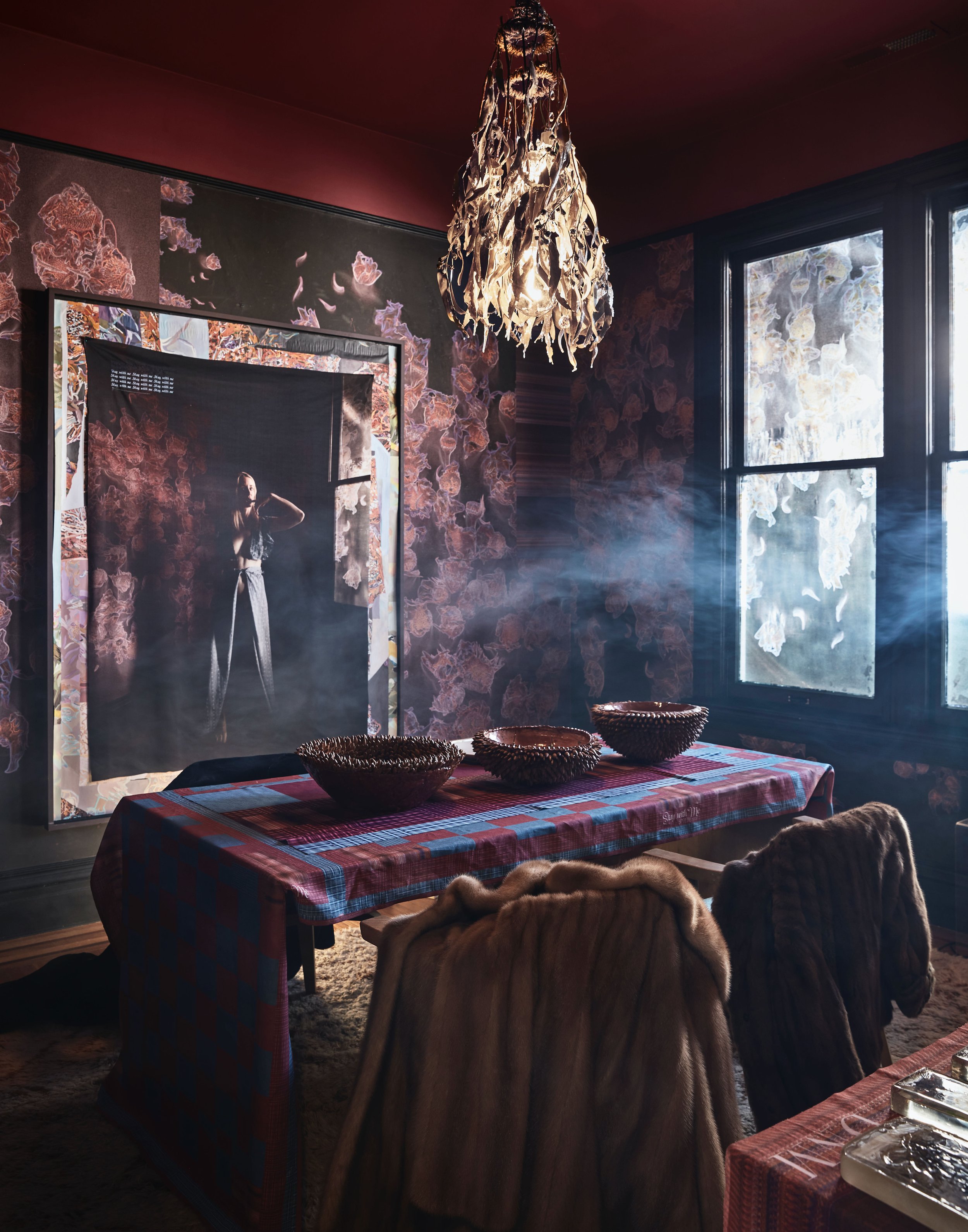 A dining room with a table covered with a red checkered tablecloth, adorned with three decorative bowls. There are fur coats draped over the chairs around the table. The room has dark floral wallpaper, large windows letting in natural light, and a ch