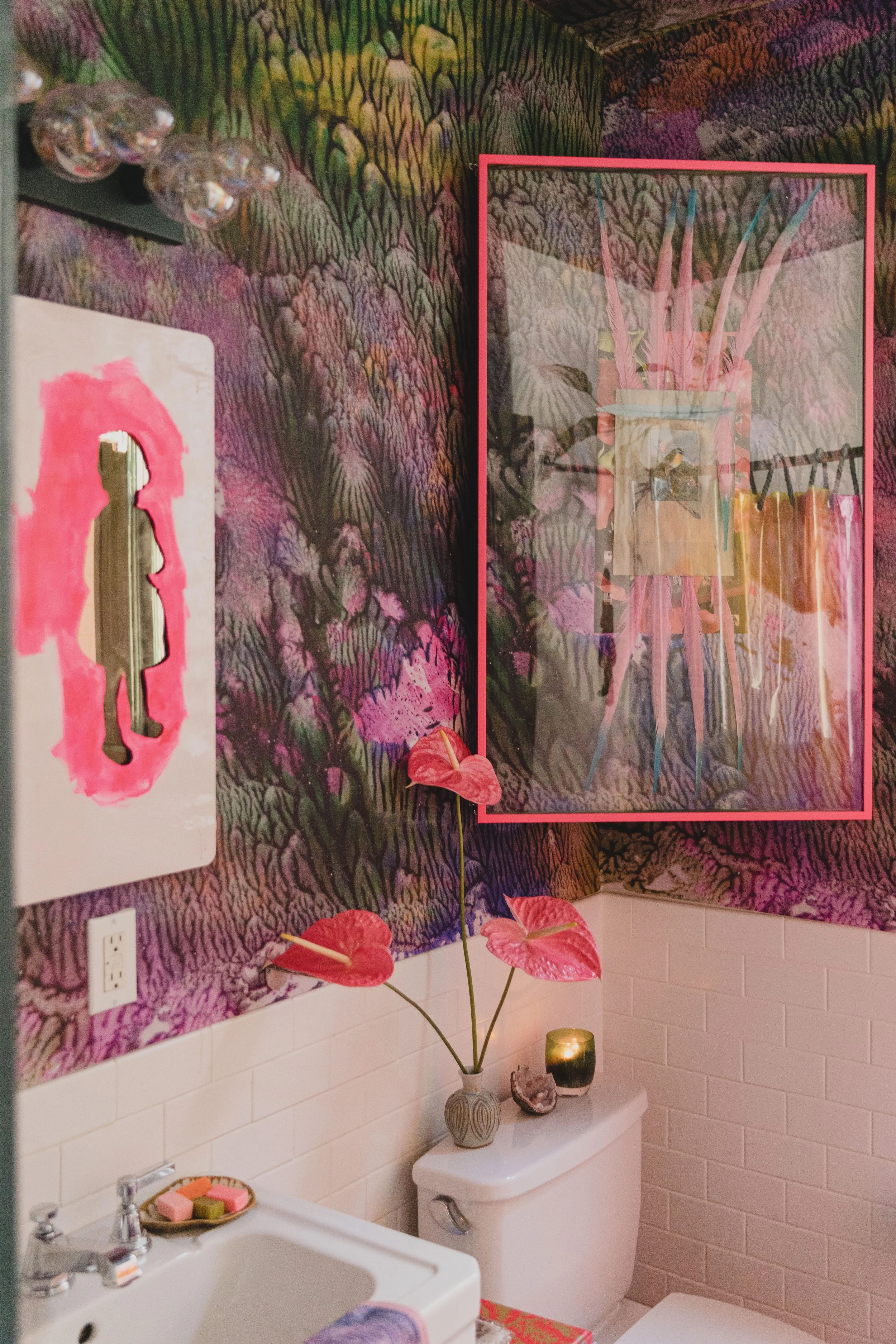 A bathroom with a vibrant, colorful wallpaper featuring a botanical design. On the wall, there's a framed mirror reflecting a plant and hanging pink fabrics. Below, a sink with soap and nearby a toilet with a decorative vase holding pink anthuriums, 