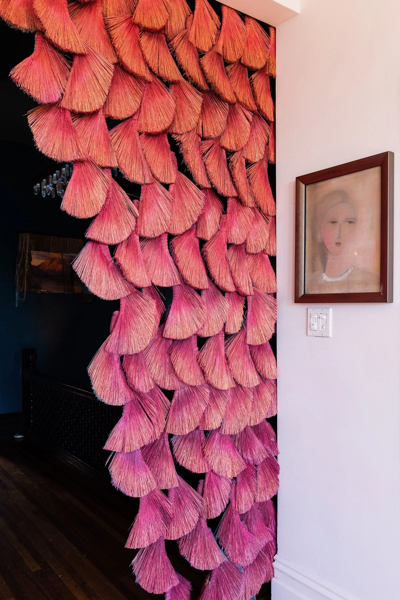 Interior of a room with a decorative wall hanging made of pink and coral-colored tassels arranged in a cascading pattern next to a framed portrait of a woman on a white wall.