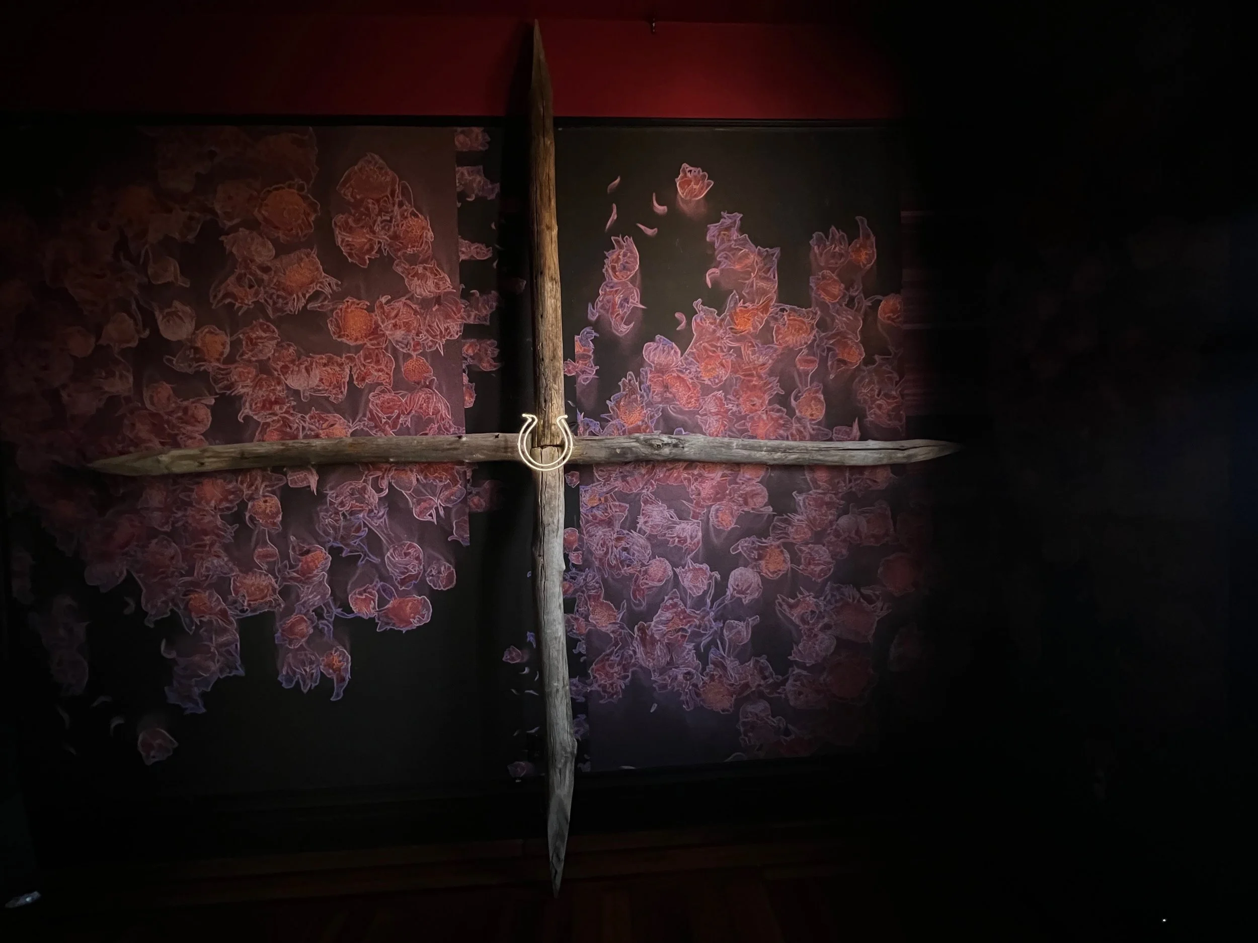 A large wooden cross mounted on a wall with a floral background, in a dimly lit setting.