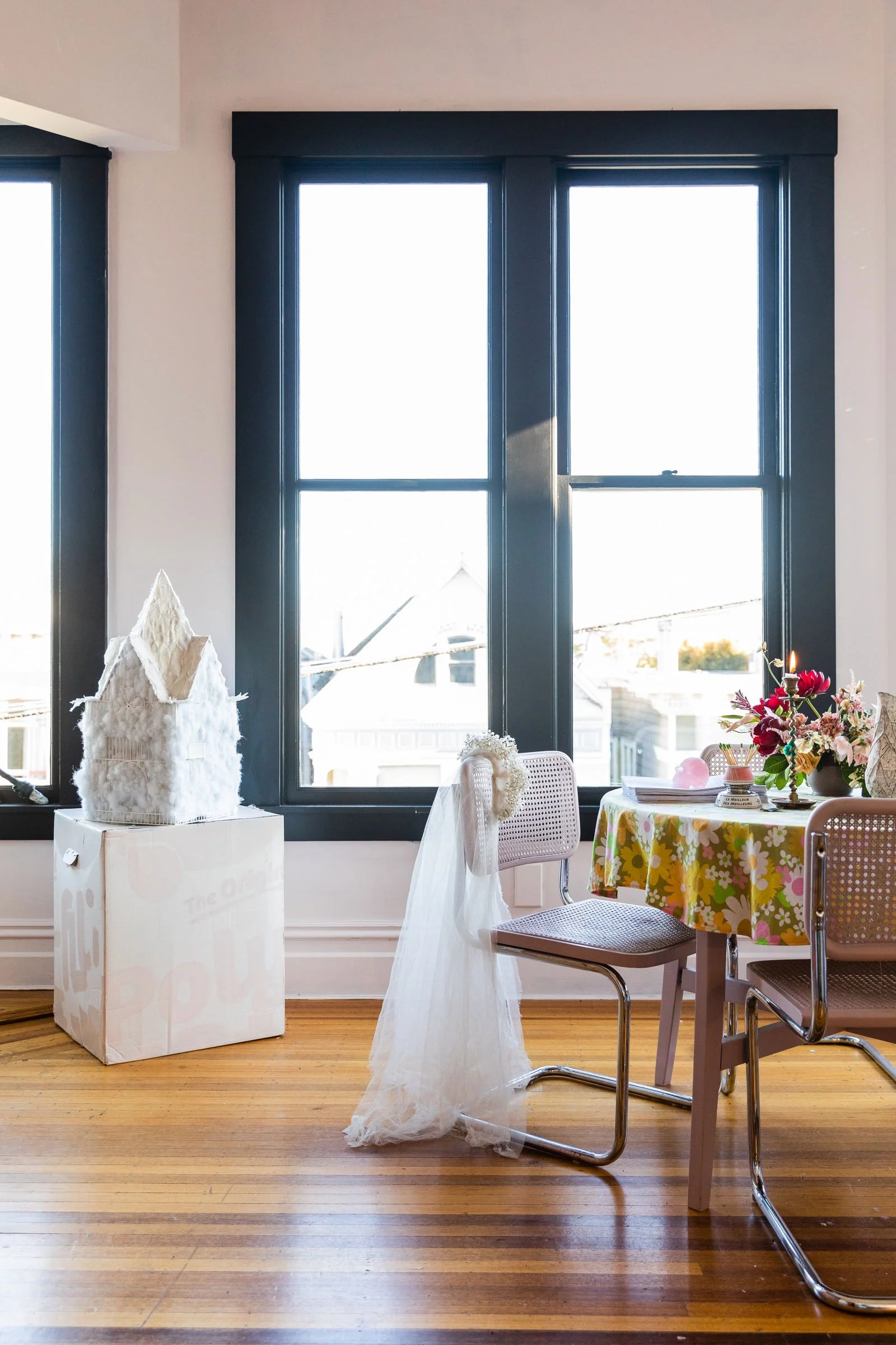 A room with large black-framed windows, a table with a floral tablecloth, a floral arrangement, and chairs, one decorated with a bridal veil and headpiece, and a white decorative house on a box.