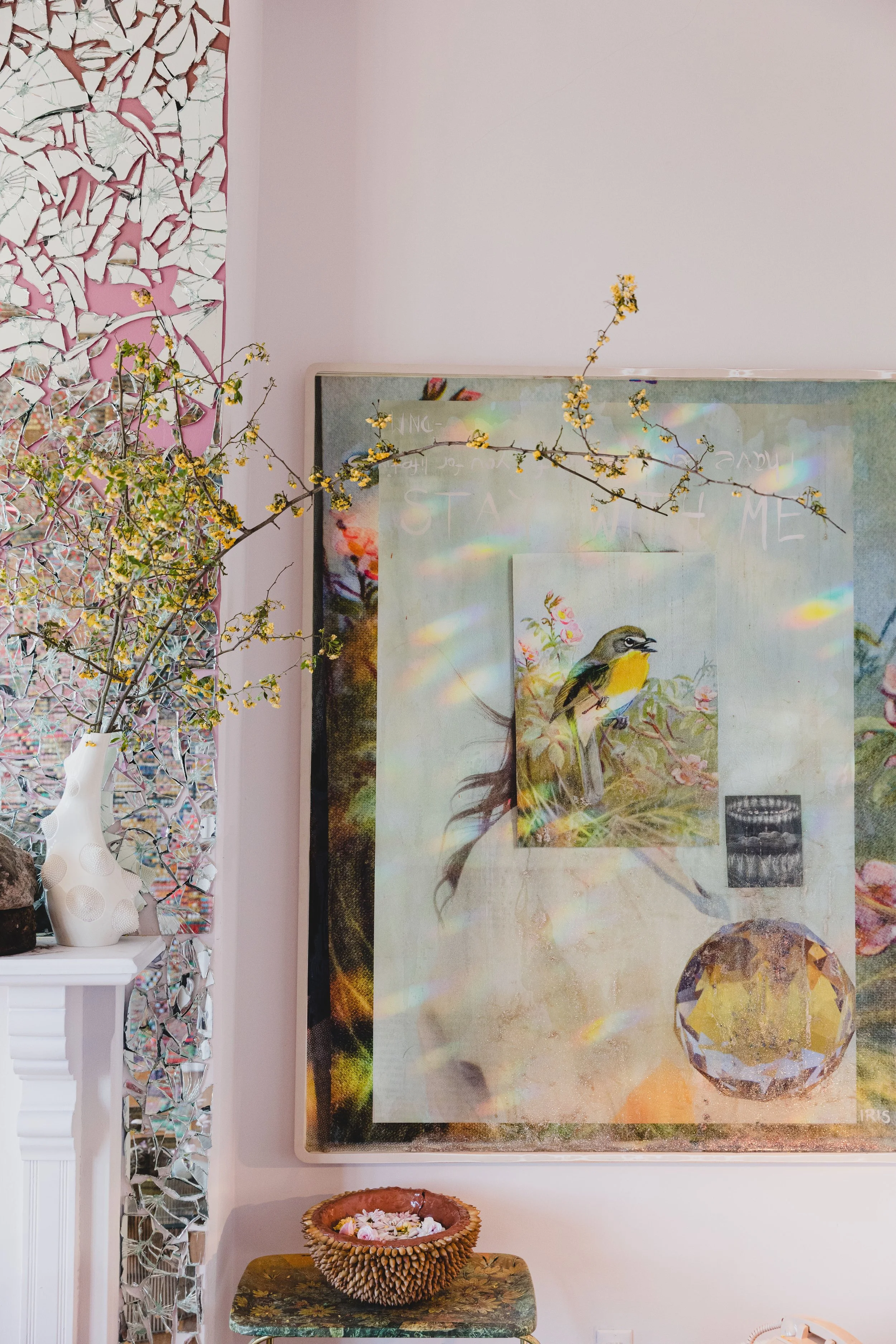 Decorative interior with a large artwork featuring a bird, yellow flowers, and colorful reflections; a white vase with branches and flowers on a white mantel; a small woven bowl with flowers on a small table.