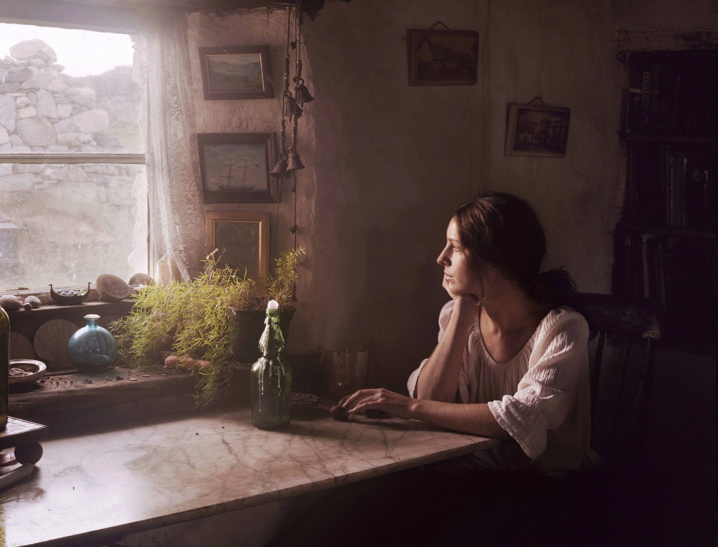 A woman with dark hair sits at a marble table, gazing out a window with lace curtains, in a cozy, rustic room with artwork and potted plants.
