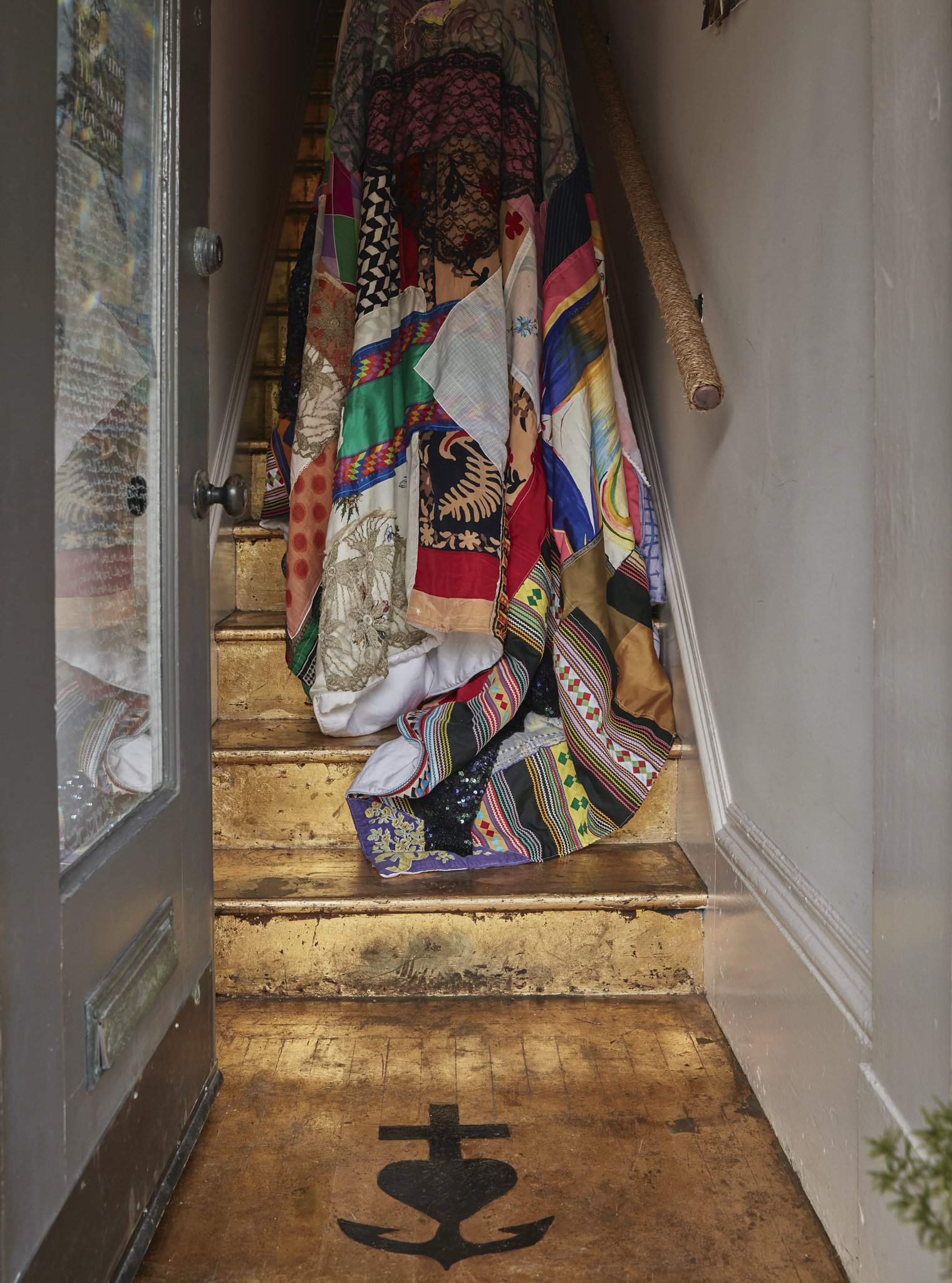 Colorful quilt draped over a staircase at an entryway, with a black anchor symbol on the wooden floor and an open door to the left.