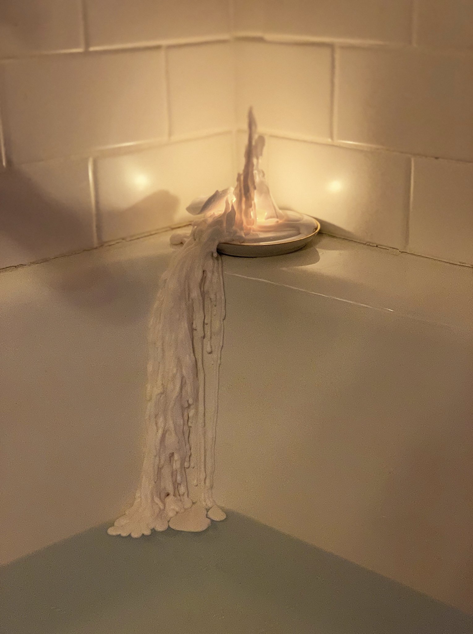 A candle on a ceramic dish has melted, with wax dripping down and pooling at the edge, creating a wax cascade against a tiled wall.