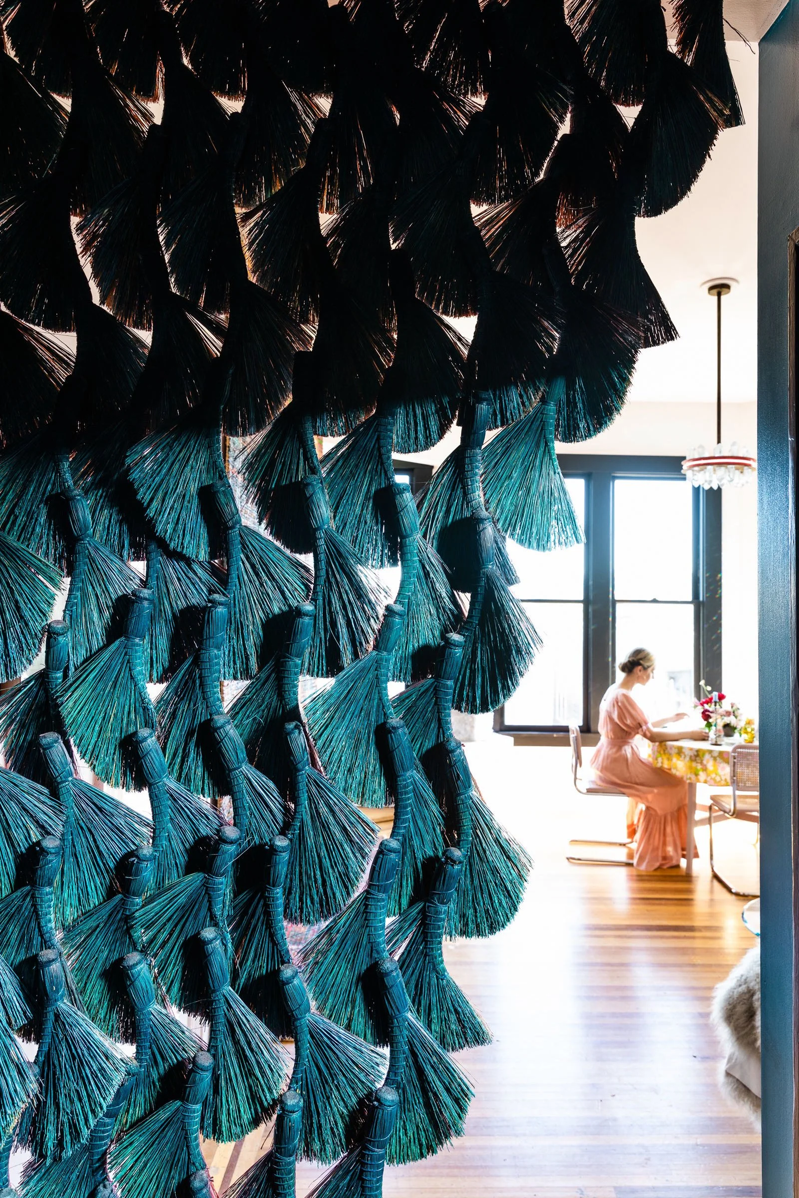 A decorative wall of black straw tassels with teal accents dividing a bright room where a woman in a pink dress sits at a floral table, near large windows letting in natural light.