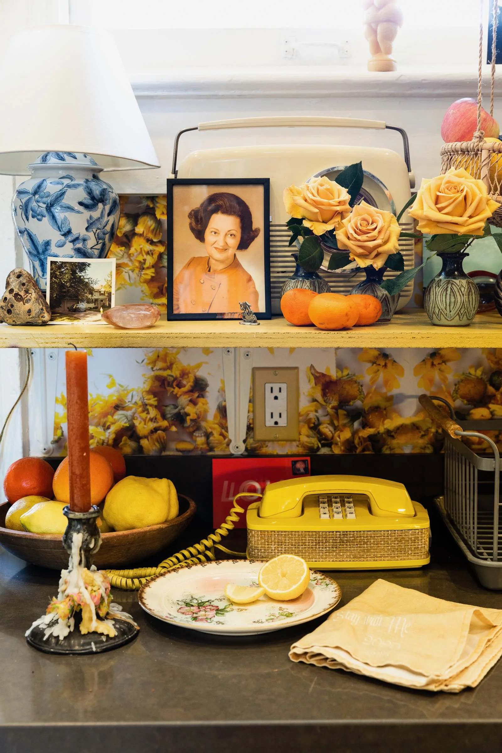 Sundry kitchen countertop items with a yellow rotary phone, a lemon half on a floral plate, a bowl of apples and citrus, a candle, decorative flowers in vases, a framed photo, a retro radio, and a vintage yellow light switch.