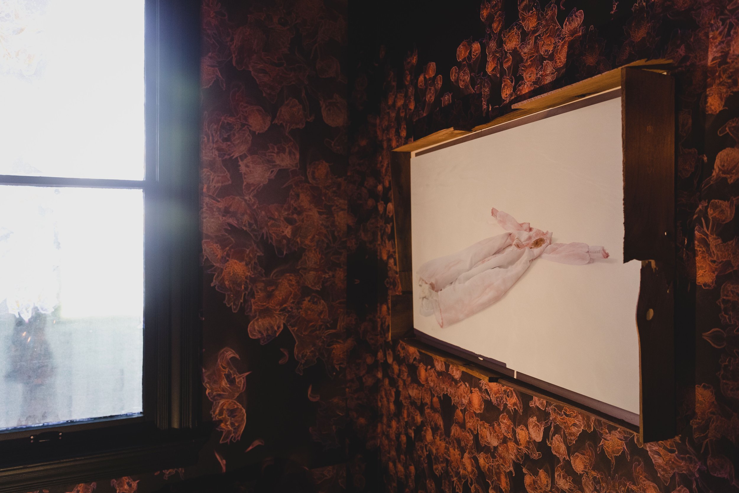 A raw chicken on a white cutting board inside a room with floral wallpaper and a window in the background.