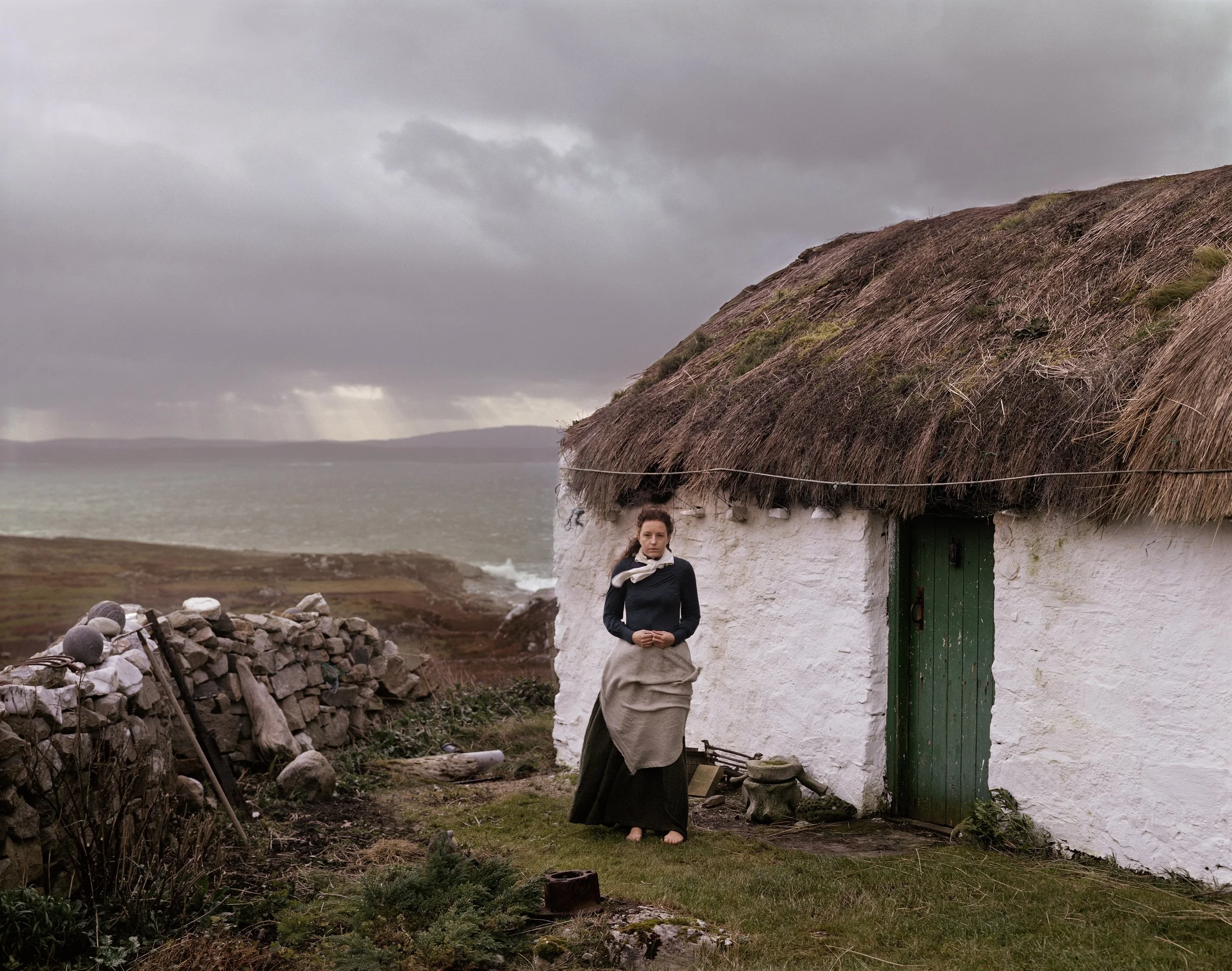 A woman standing outside a small white house with a thatched roof, near the coast under a cloudy sky.