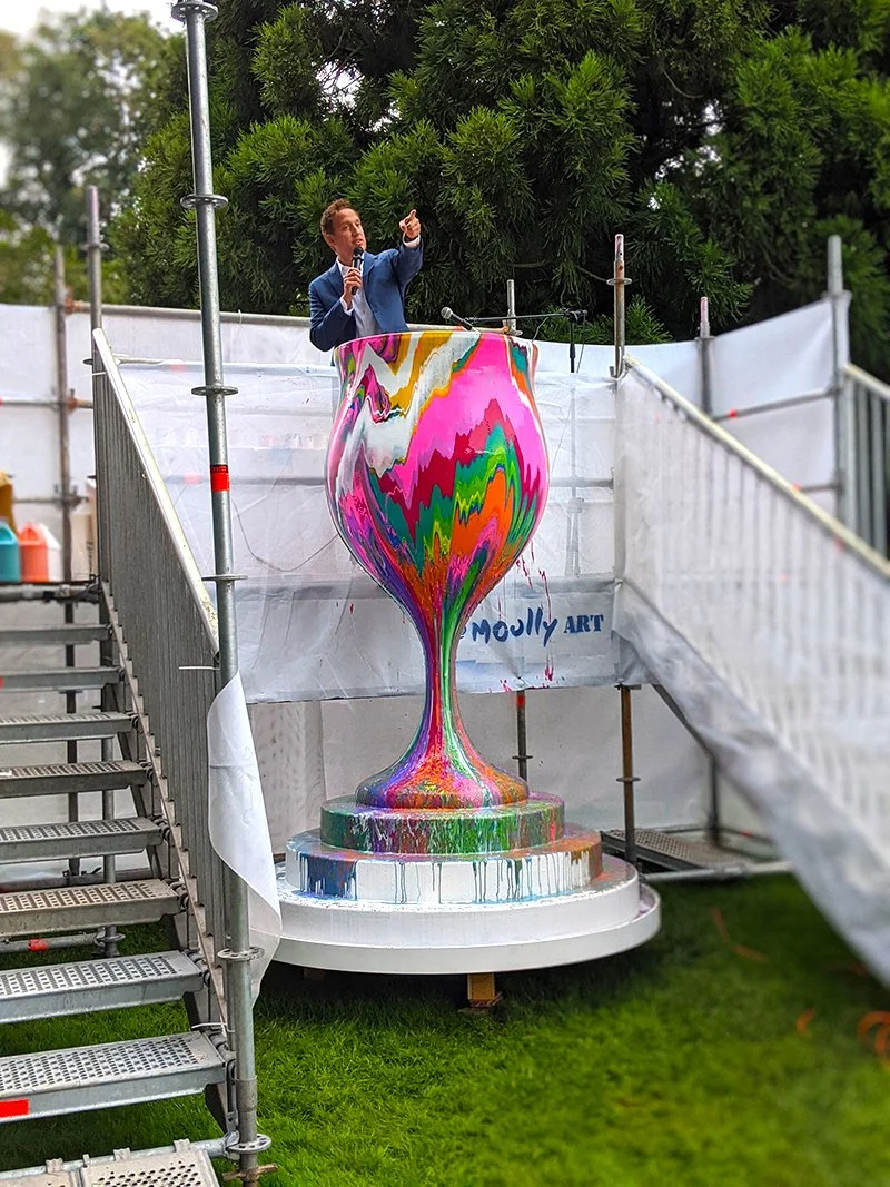 Man in a blue suit speaking into a microphone on a platform next to a large, colorful, abstract sculpture resembling a vase or goblet, outdoors at an art event.