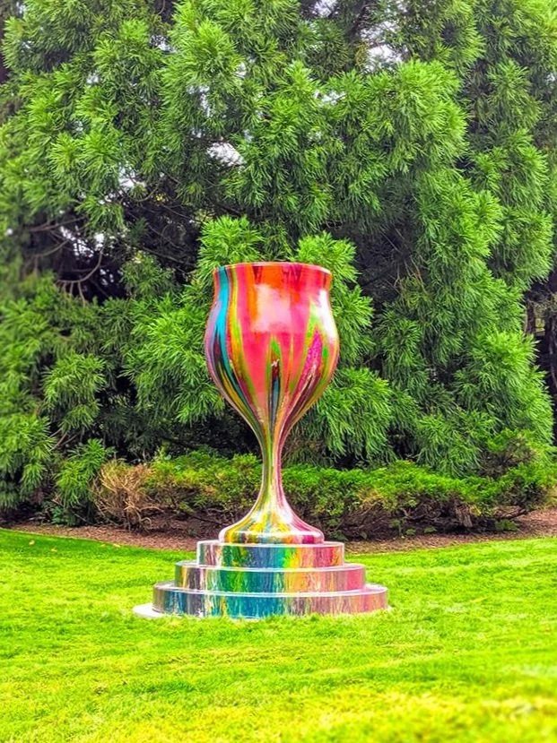 Large multicolored vase sculpture with a drip paint effect, placed on a stepped pedestal, outdoors on a grassy field with trees in the background.