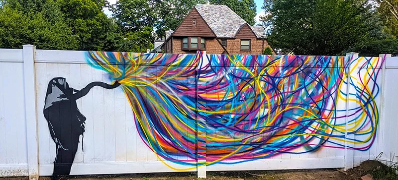 A mural on a white fence depicting a silhouette of a person with colorful, flowing lines extending from their head, symbolizing thoughts or creativity.