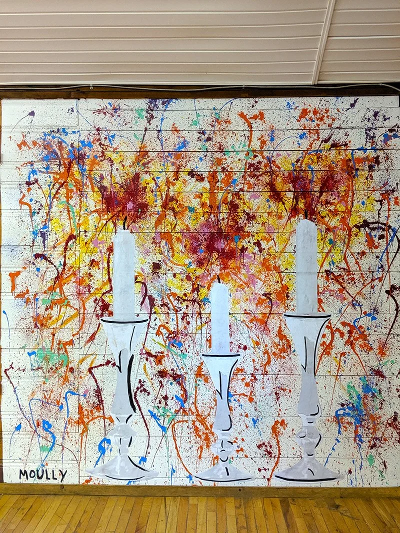 Colorful abstract mural featuring three white candle holders with candles and splattered paint in red, yellow, blue, purple, and green on a white brick wall.