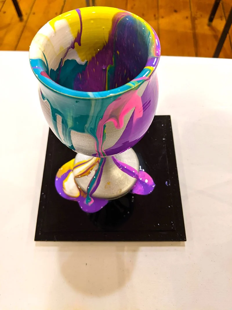 A ceramic goblet with colorful paint drips, placed on a black square base on a white surface.