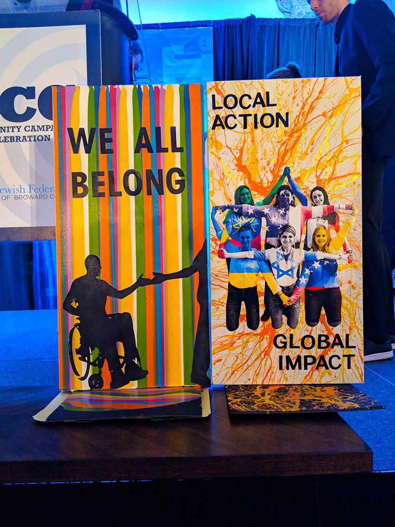 Two colorful posters on a table, one with the message 'WE ALL BELONG' and an illustration of a person in a wheelchair sharing a handshake, the other with the message 'LOCAL ACTION, GLOBAL IMPACT' and a group of diverse people holding hands with artis