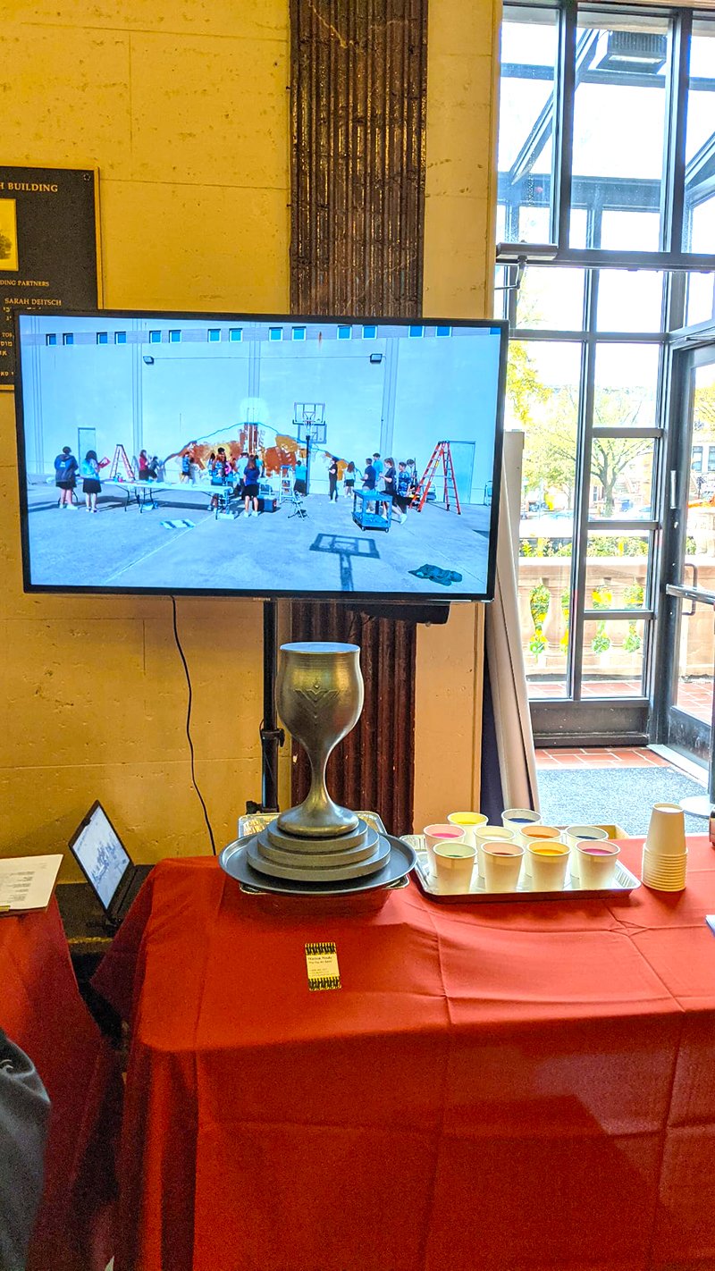 A table with a large trophy, cups with colored drink, and a tray of cups, in front of a large television screen showing kids playing basketball in a gymnasium.