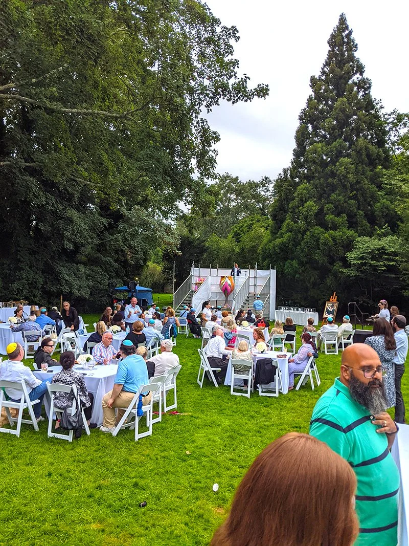 People gathered at an outdoor event on a grassy lawn with trees in the background, sitting at round tables with white chairs, watching a stage with performers and a large pink inflatable unicorn.