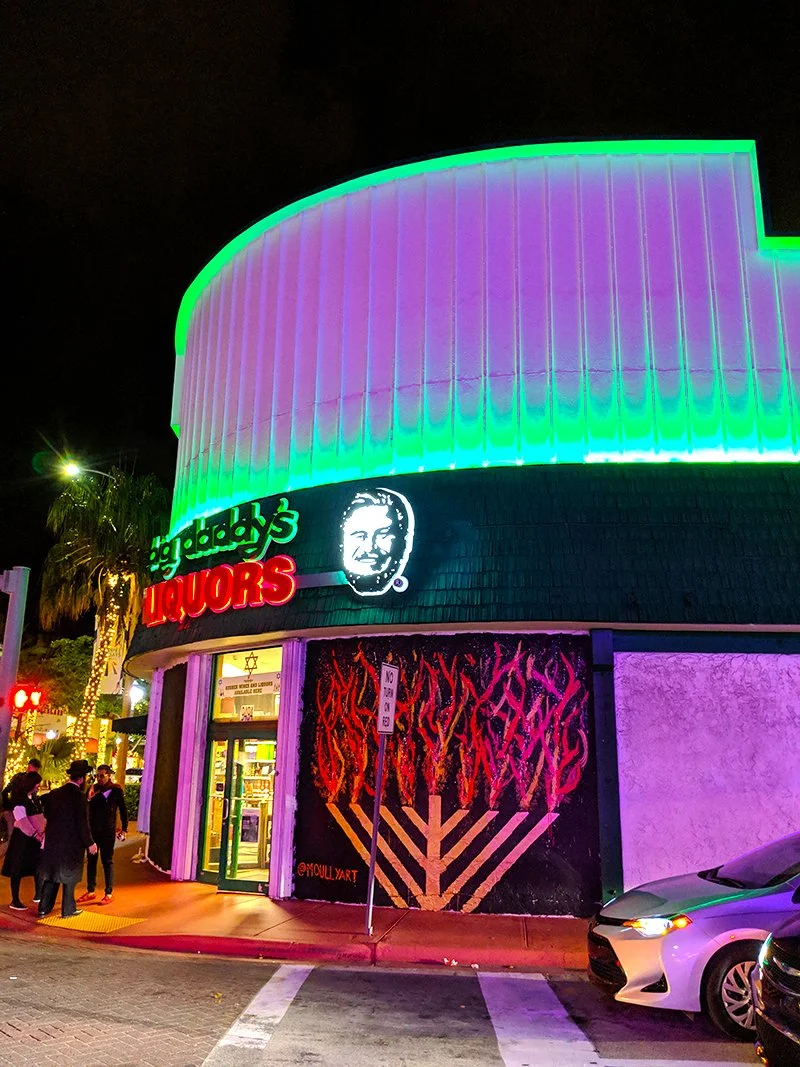 Nighttime street scene featuring a brightly lit building with neon lights, a sign for Gregory's Liquors, and a mural with a menorah and flames, with people gathered outside and parked cars.