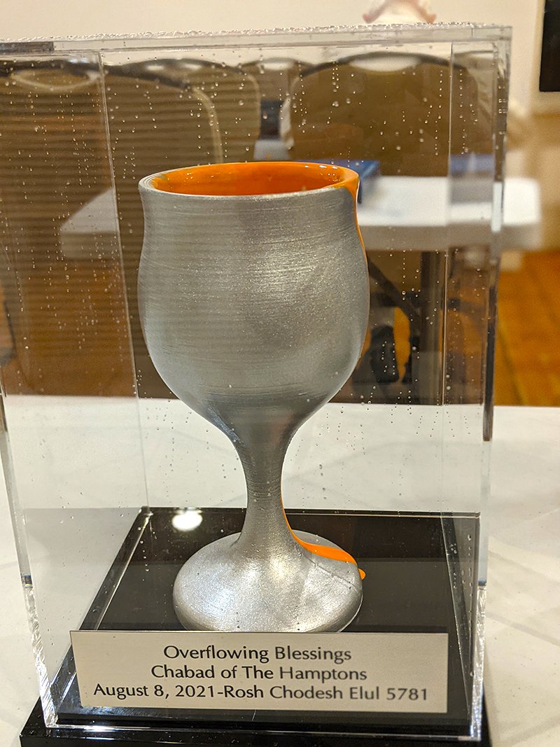 A silver goblet with an orange interior, displayed in a clear case. A plaque beneath the goblet reads "Overflowing Blessings, Chabad of The Hamptons, August 8, 2021-Rosh Chodesh Elul 5781."
