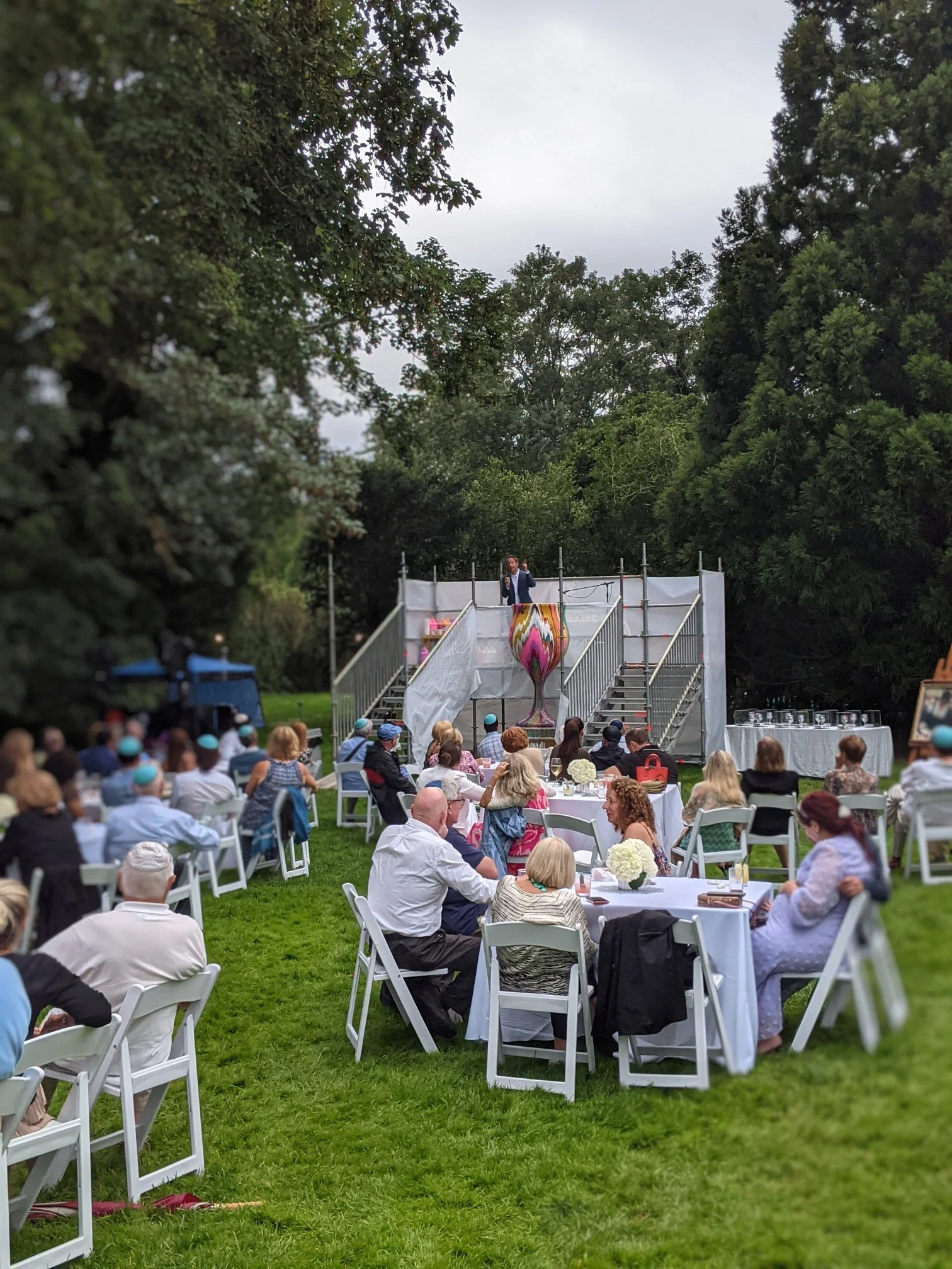 Outdoor Jewish community celebration featuring Yitzchok Moully public art — garden ceremony with guests seated at tables and speaker at stage
