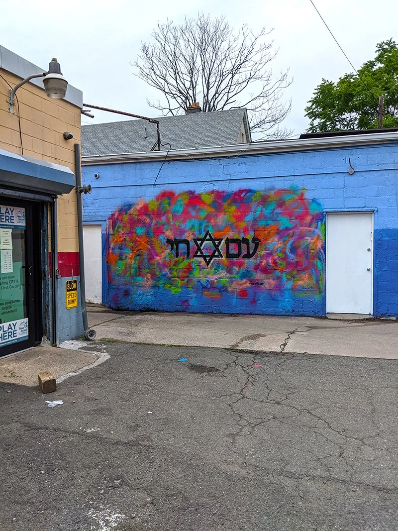 Colorful graffiti mural on a blue wall with Hebrew letters and a Star of David, next to a street scene with cracked pavement and a storefront.