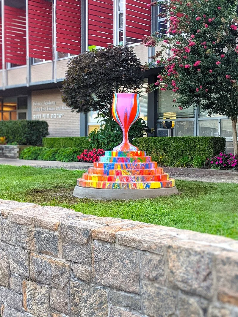 Colorful sculpture of a large goblet on top of a stepped pedestal, placed in a garden with green grass, trees, and bushes, near a modern building with red and gray exterior.