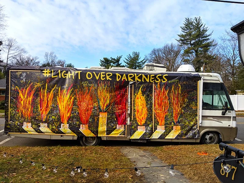 A large truck with a painted mural of seven burning candles and the hashtag #LightOverDarkness at the top. The background shows trees and a partly cloudy sky.